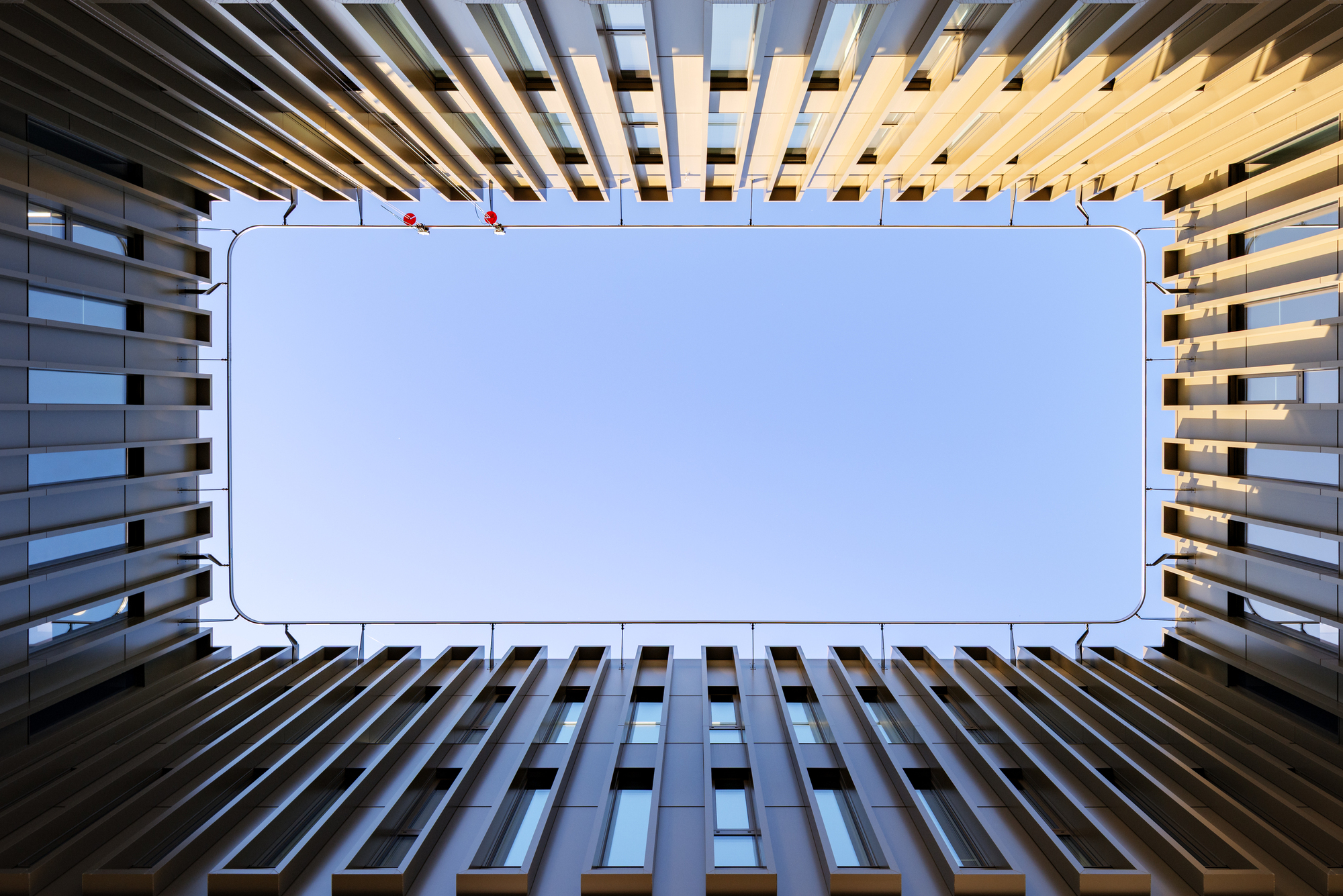 Upward view from inside the courtyard through angled vertical fins to a rectangular patch of pale sky, with warm sun on the edges
