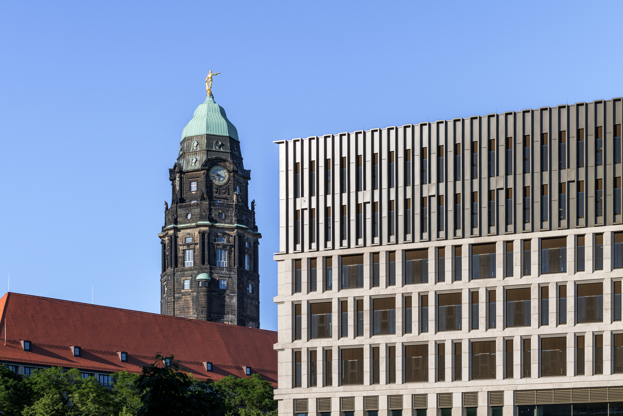 Gridded stone facade with finned upper floors beside the old clock tower and red tiled roof, under a clear blue sky