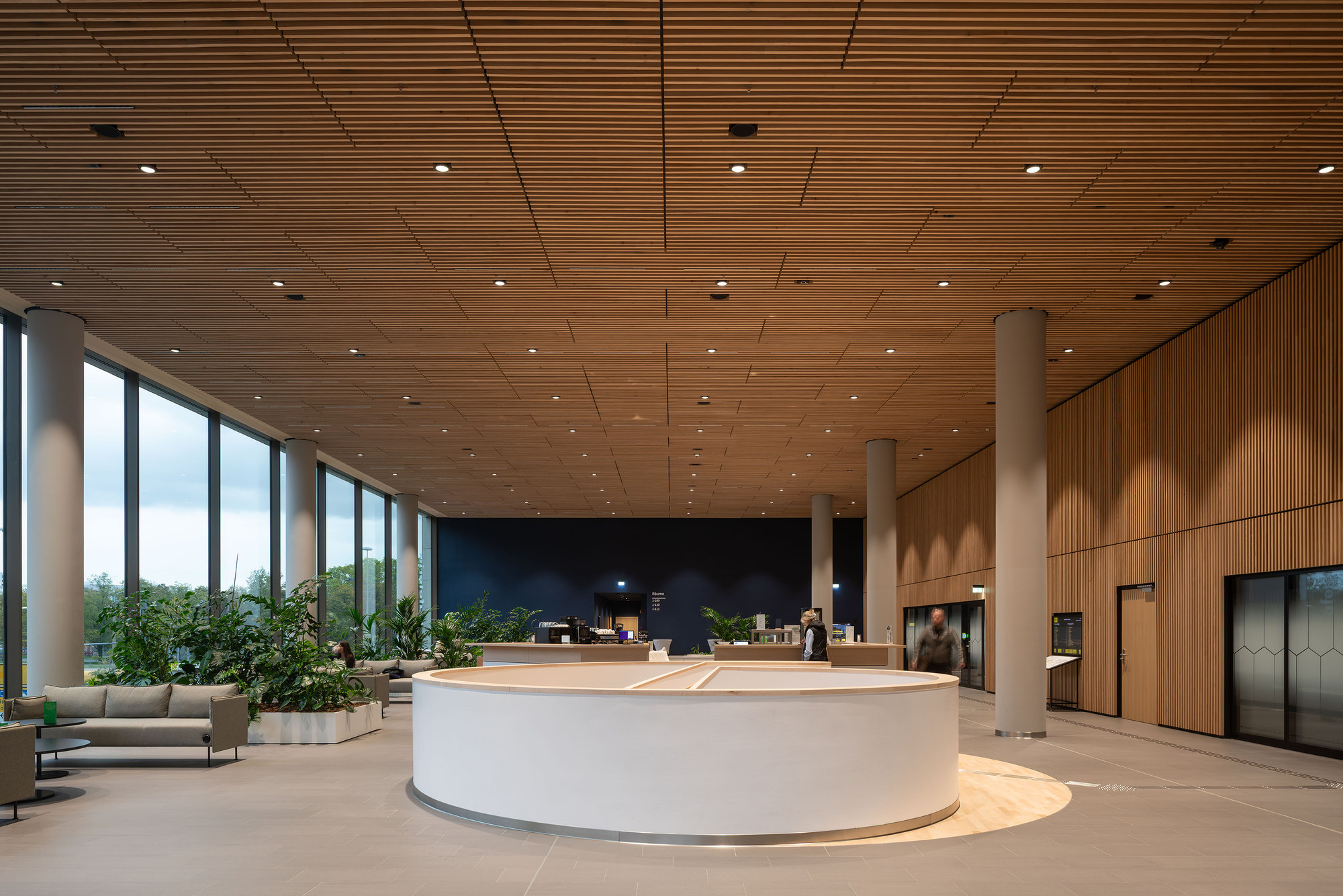 Circular white reception desk under a timber slat ceiling, with potted plants along the glazed wall and two people behind the counter