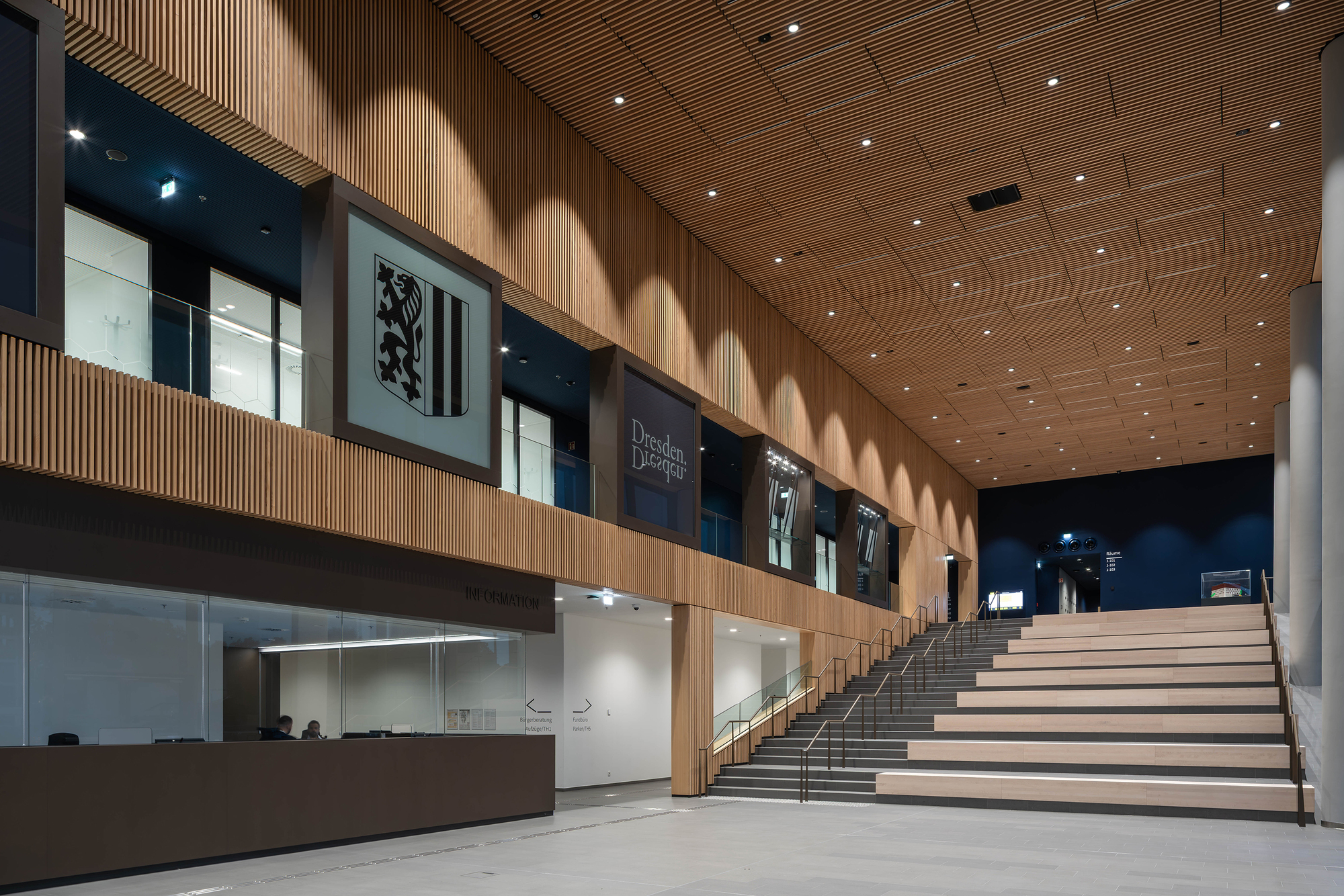 Wide timber-clad stepped seating beside a service counter, with framed crest and signage panels on the upper balcony lit at night