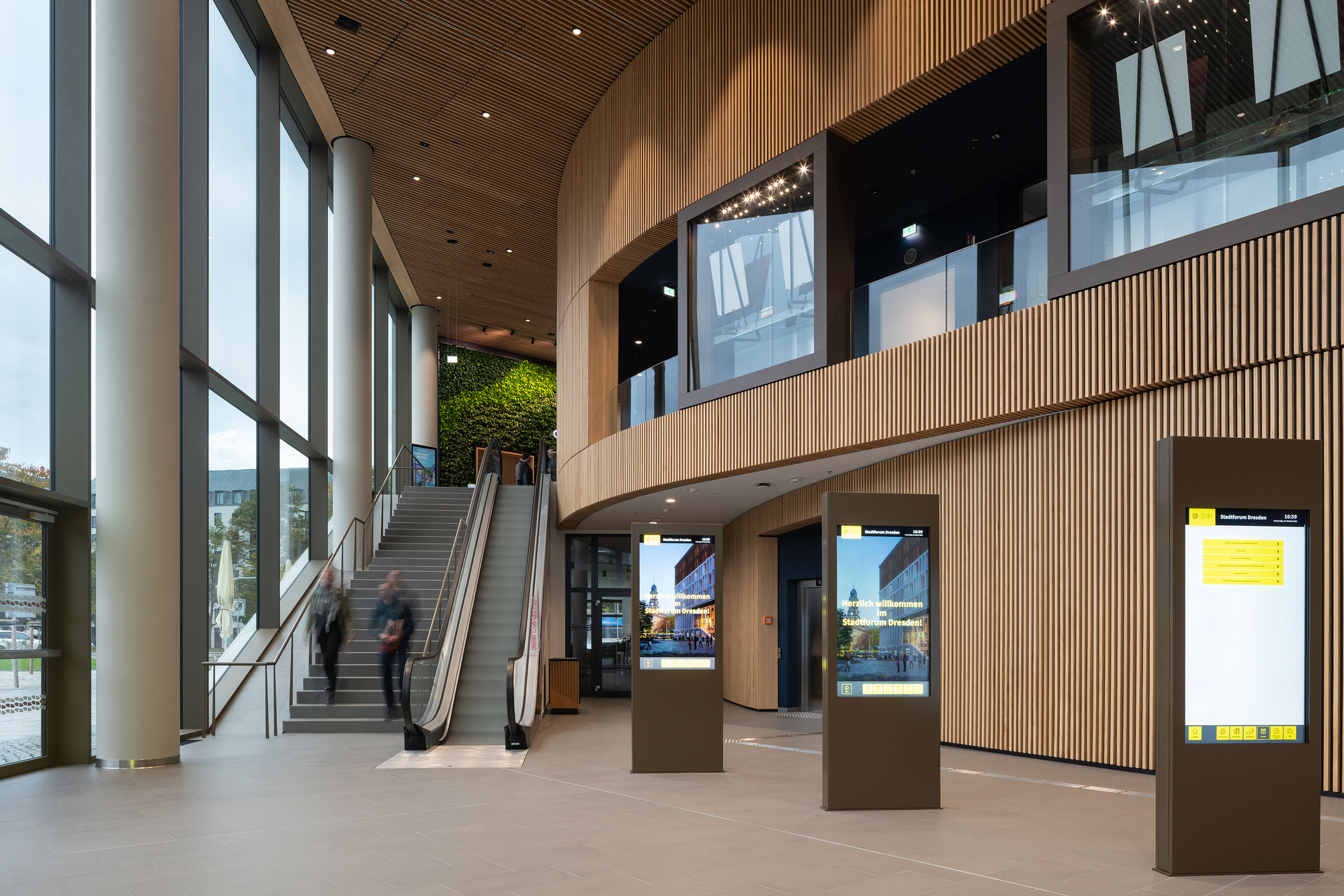 Escalator and stair beside a tall glazed wall, with three freestanding digital totems and blurred walkers in front of the curved timber balcony