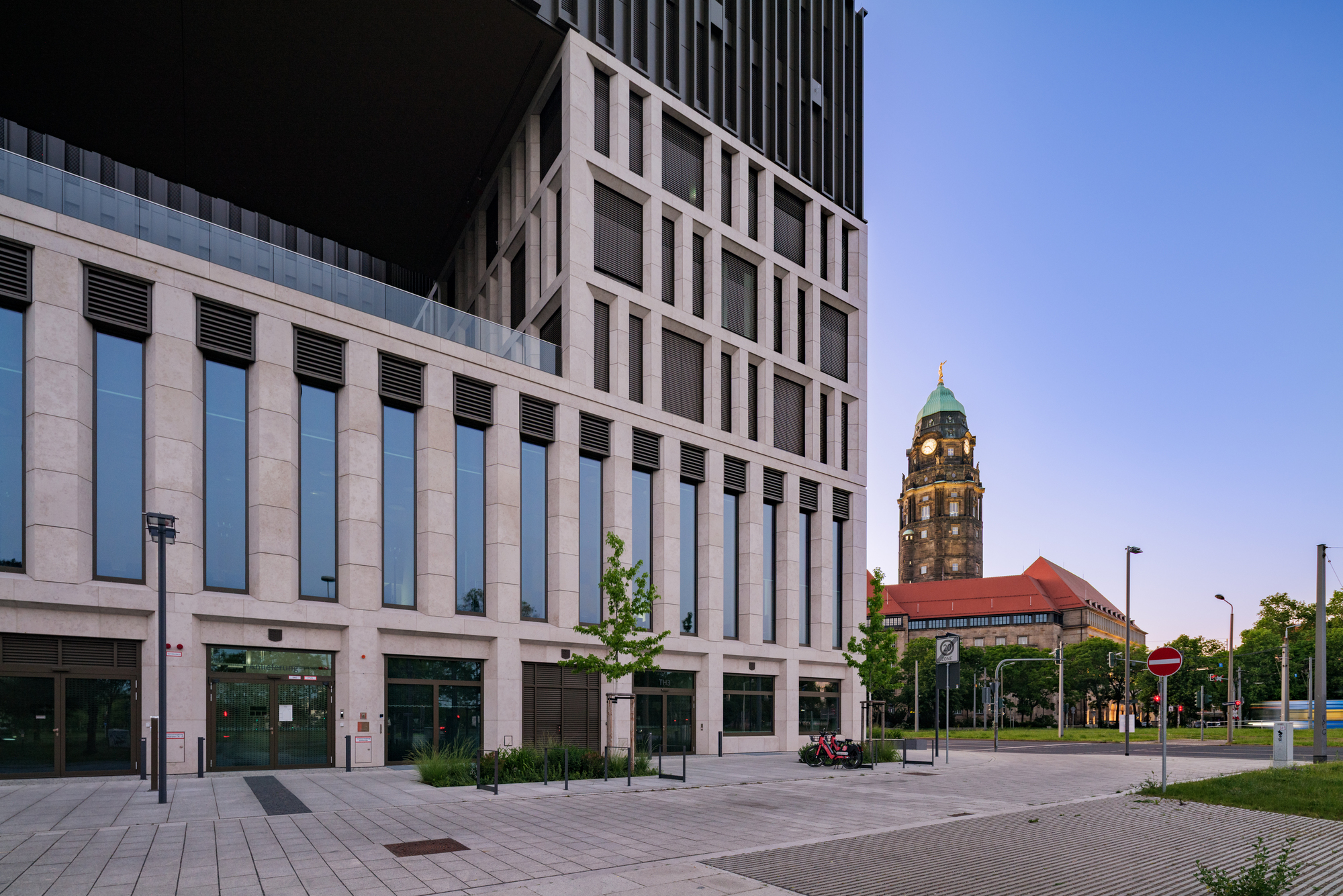 Stone facade with tall narrow windows and a young street tree, with a distant clock tower under a clear dusk sky