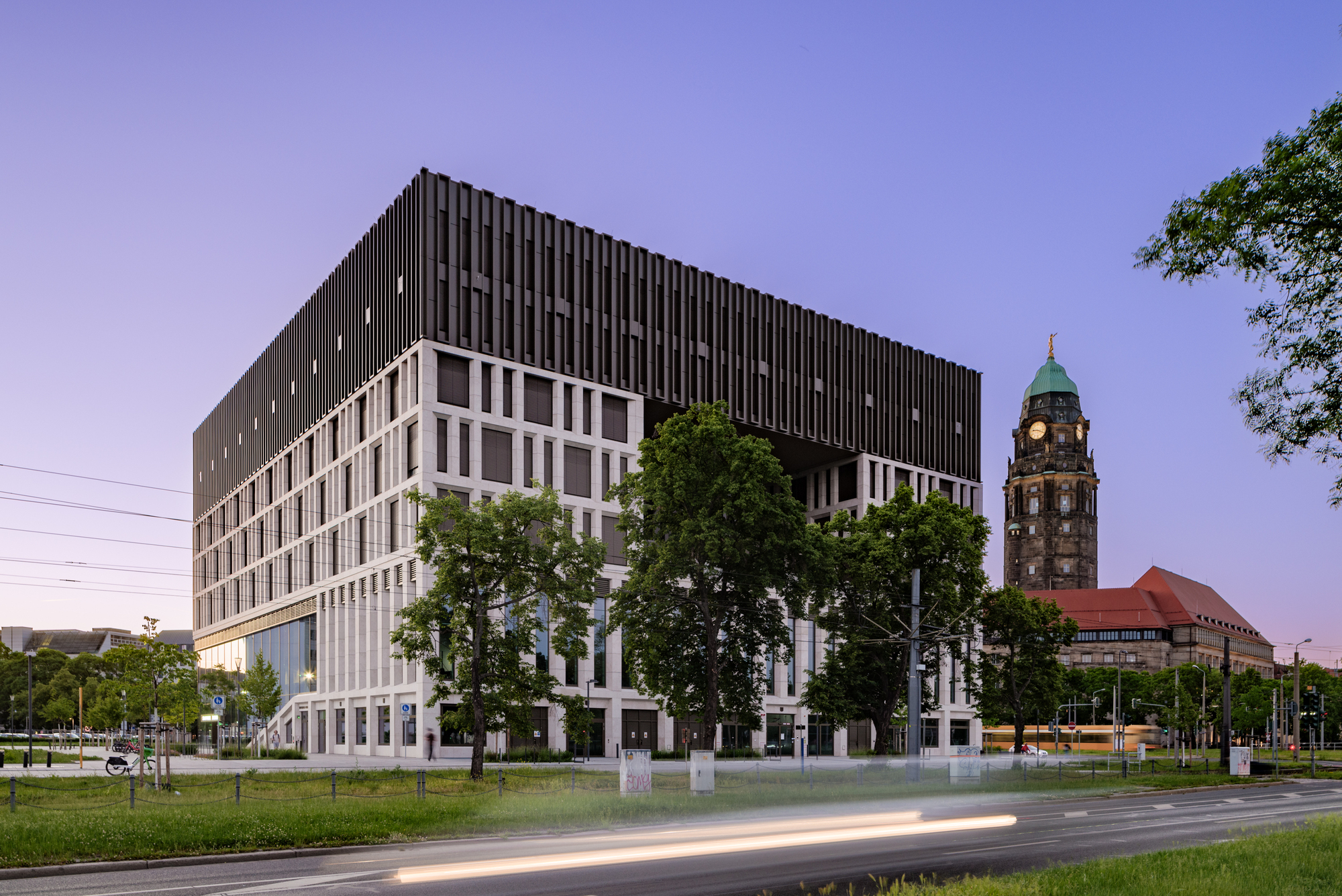 Cubic building with dark vertical fins above a pale gridded base, a clock tower behind and a light streak on the road at twilight