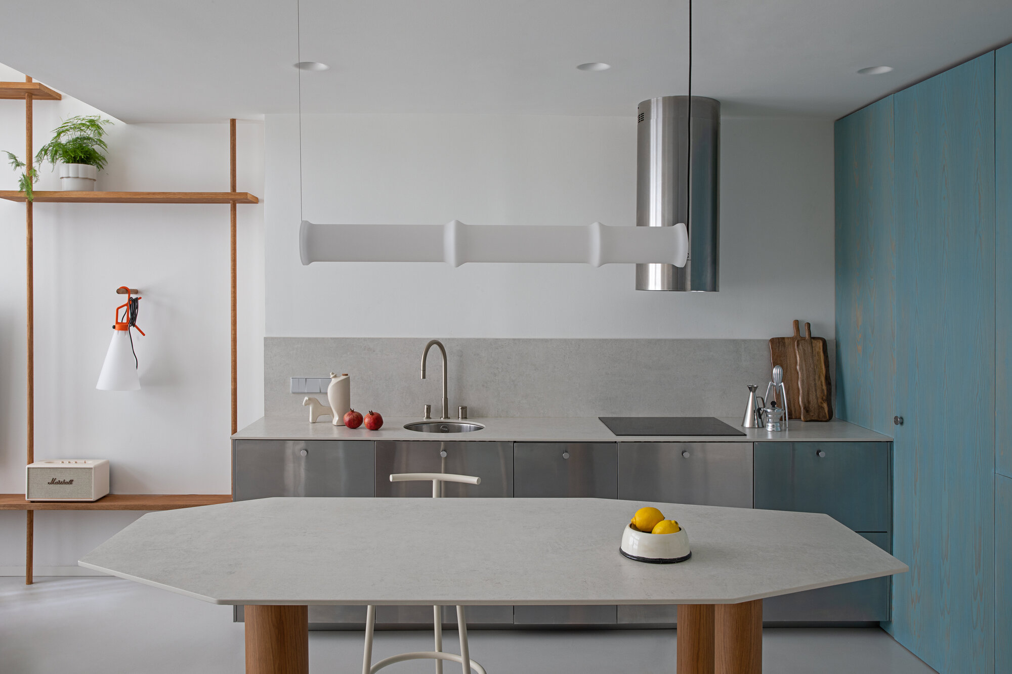 Stainless steel kitchen run with stone backsplash, faceted island table and blue wall cabinets beneath a cylindrical hood and white pendant