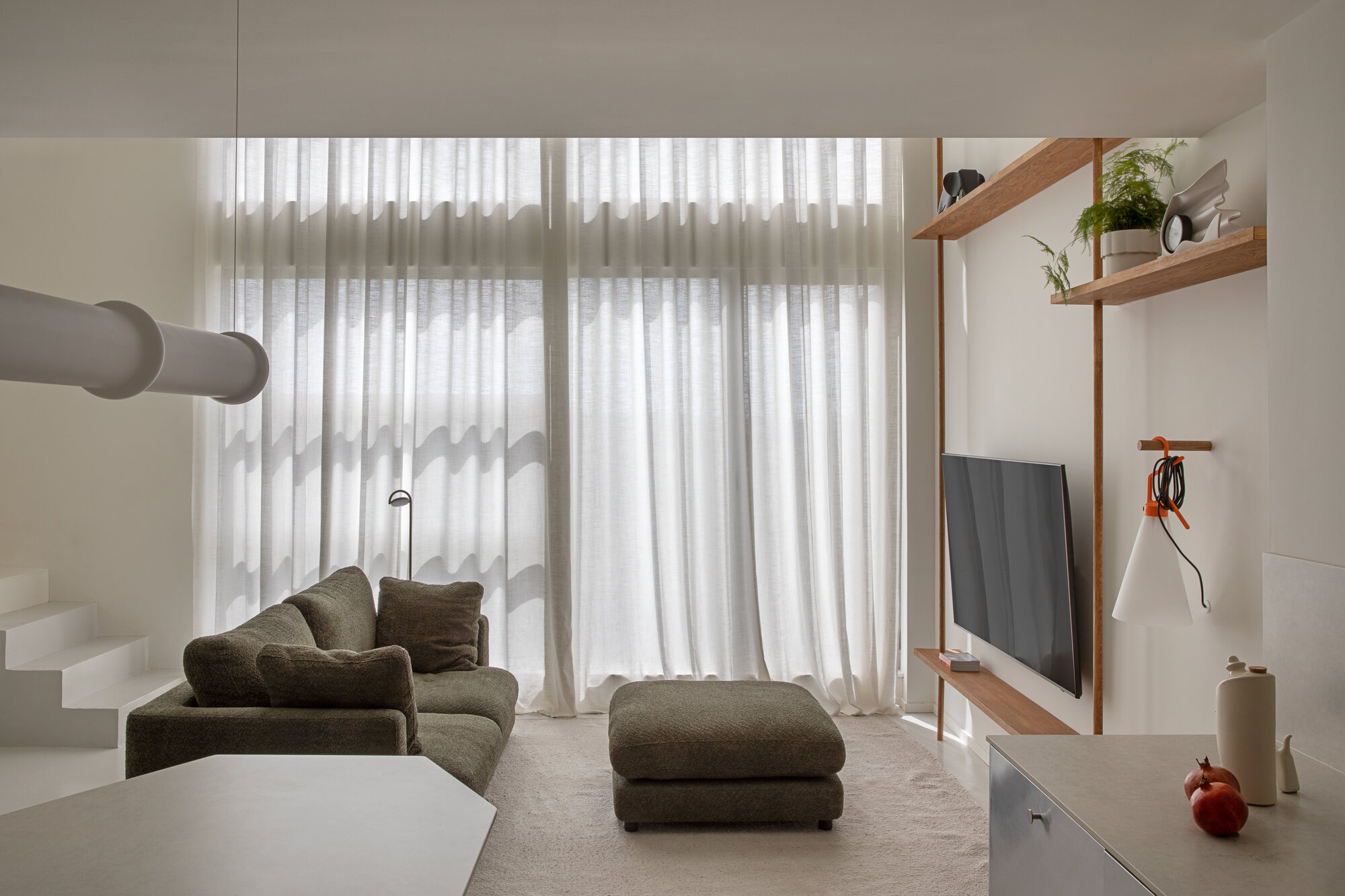 Living area with olive sofa and ottoman facing floor-to-ceiling sheer curtains that filter soft daylight, timber shelves on the right