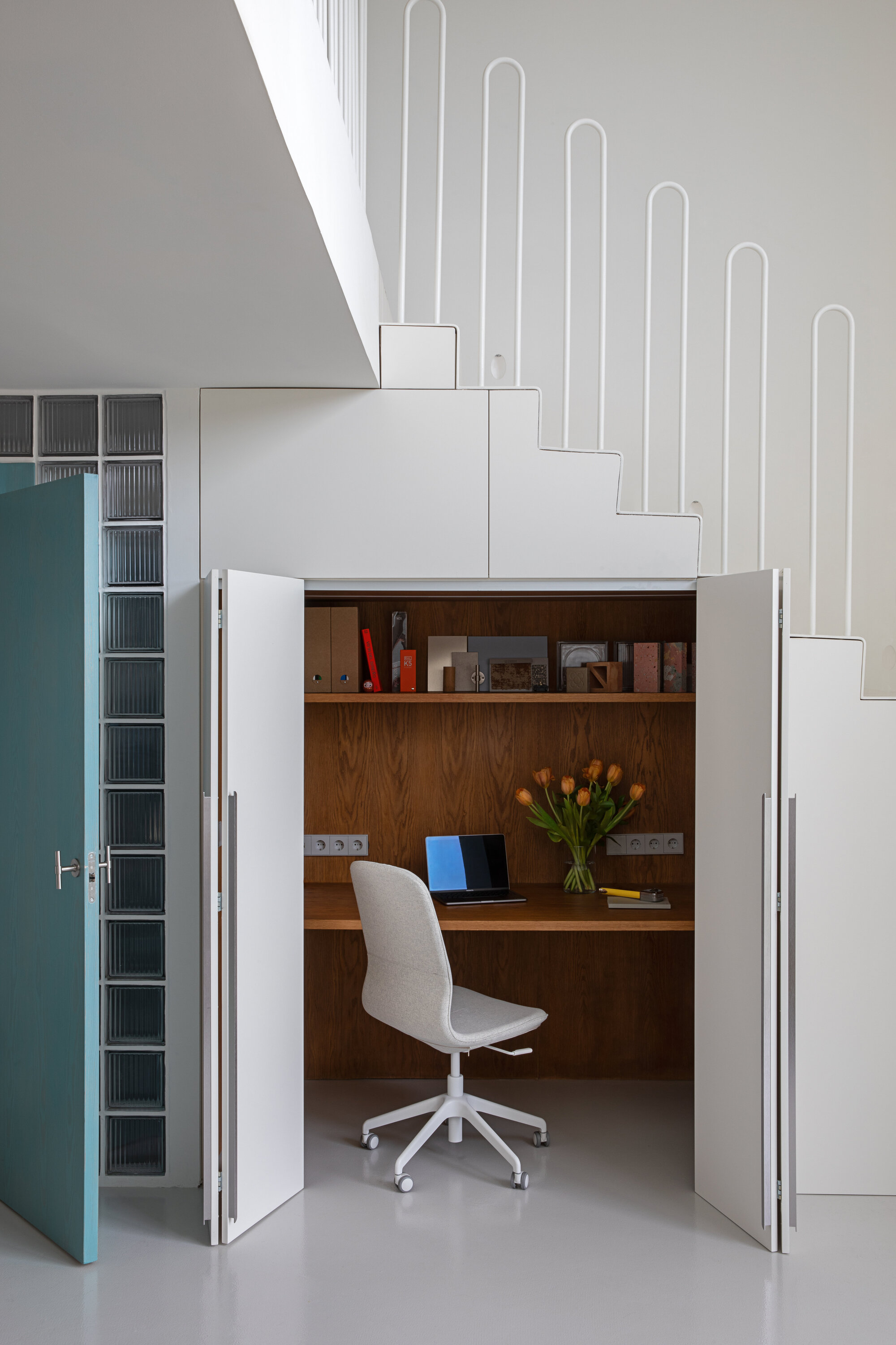 Workspace tucked under the stairs with folding white doors open on a desk, oak shelving, task chair and a vase of tulips