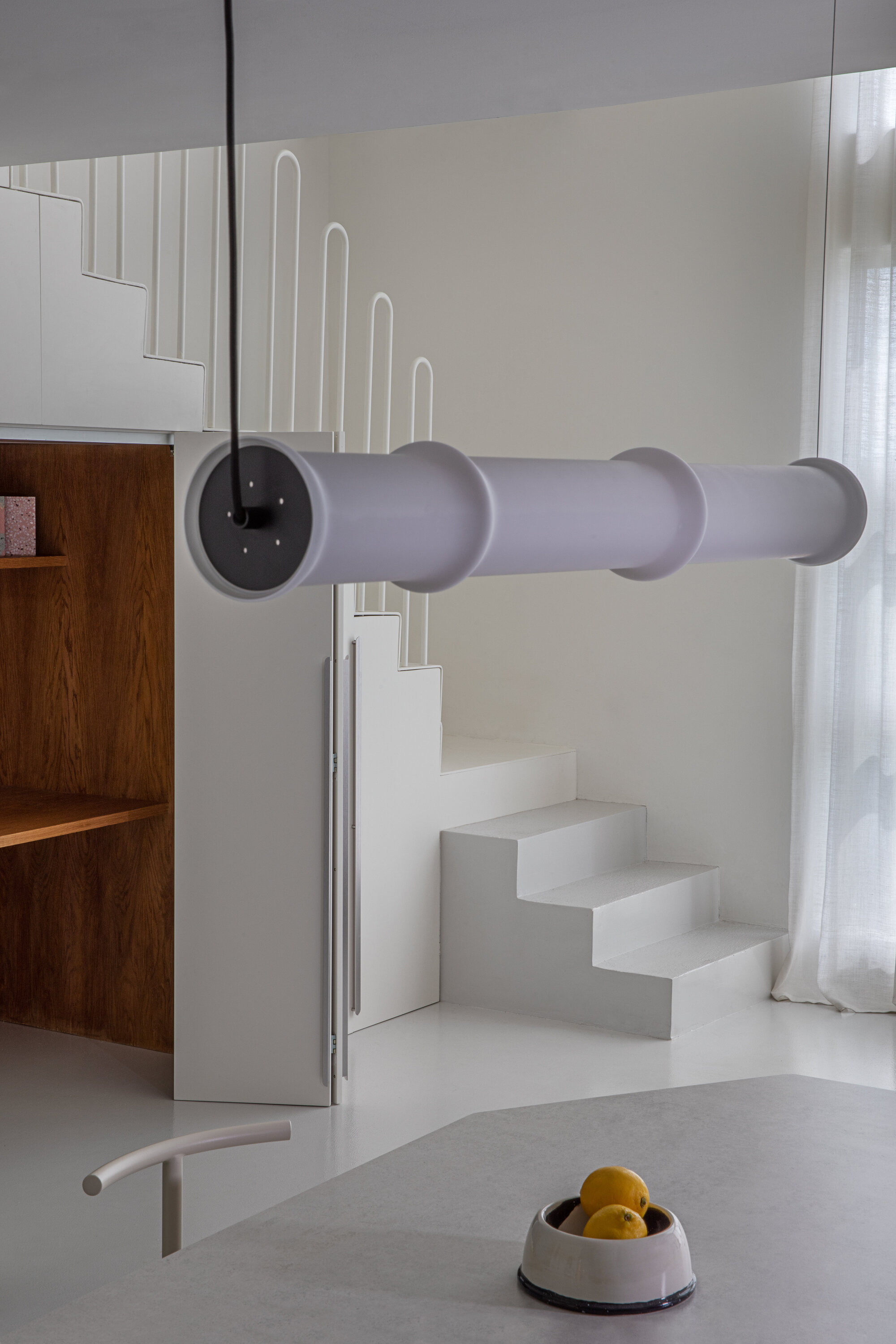 Cylindrical white pendant lamp hanging in front of the white concrete stair, with a bowl of lemons on the table below
