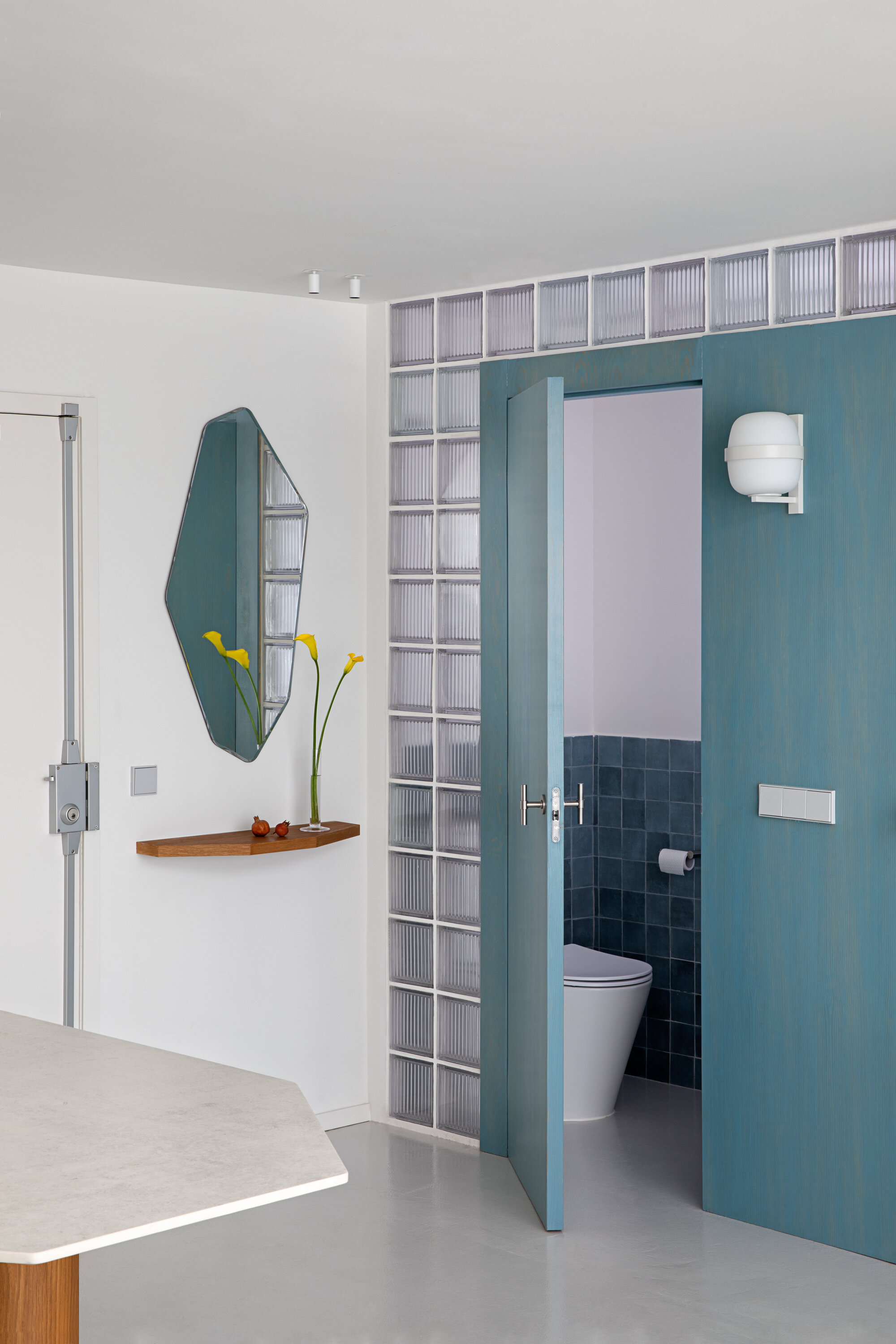 Blue timber door open to a tiled powder room, framed by a glass block wall and a faceted mirror above a wooden shelf