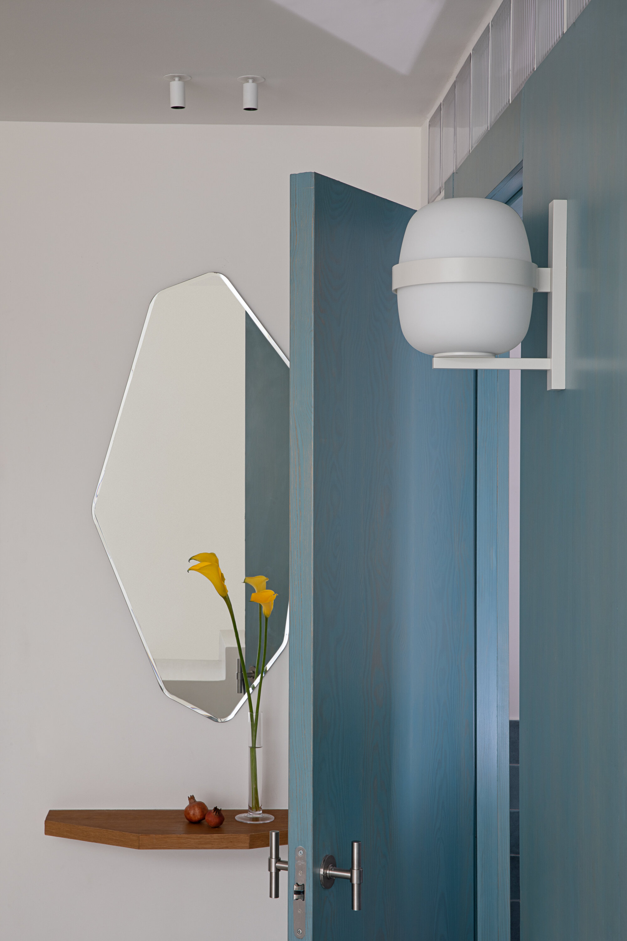 Close-up of the faceted mirror, a floating wood shelf with yellow calla lilies and a white globe wall lamp beside the open blue door