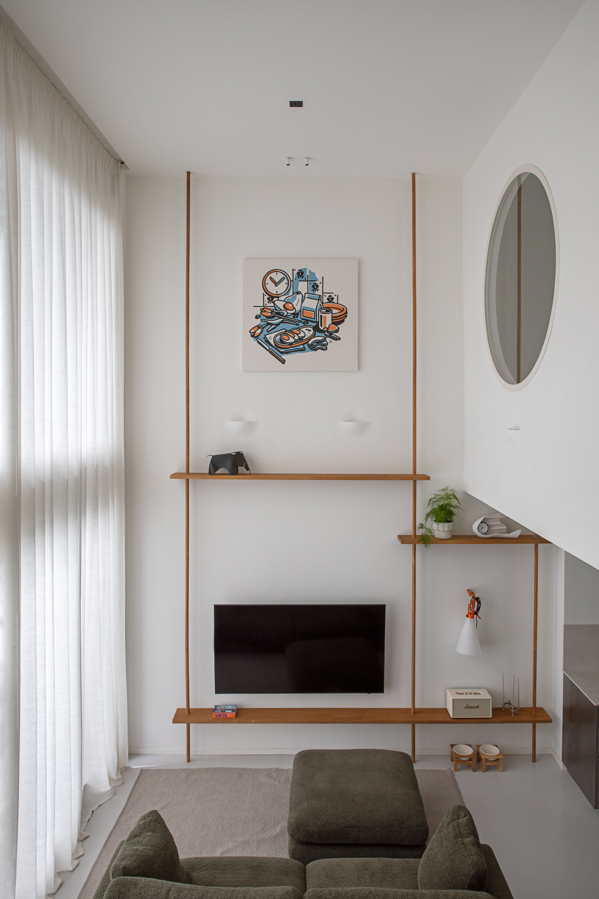 Double-height living wall with slim timber shelves hung from the ceiling, a framed artwork, a wall-mounted television and sheer curtains filtering daylight