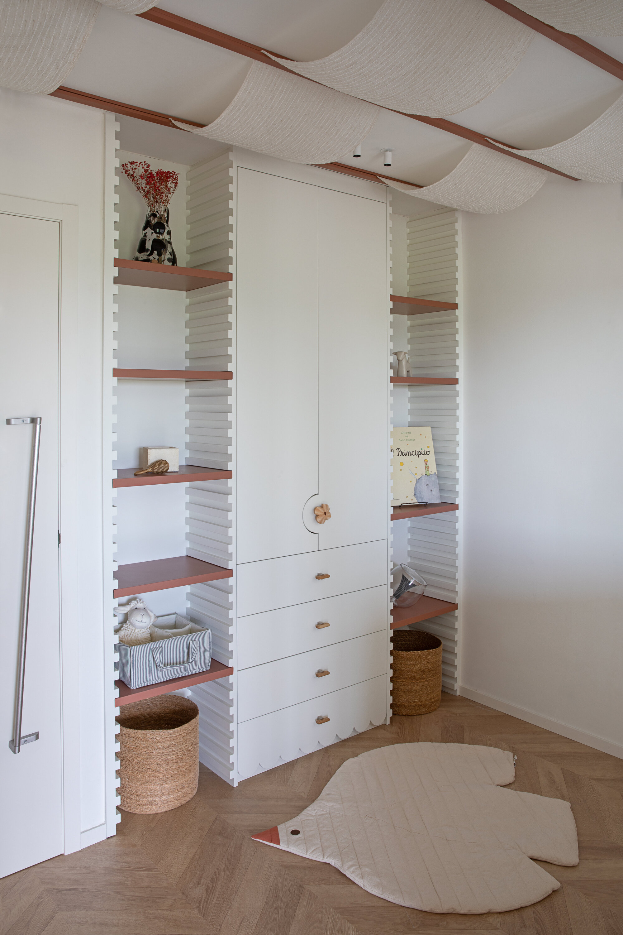 Built-in white cabinet with ribbed side panels, terracotta shelves and drawers, beneath a draped fabric ceiling and above a bird-shaped floor mat