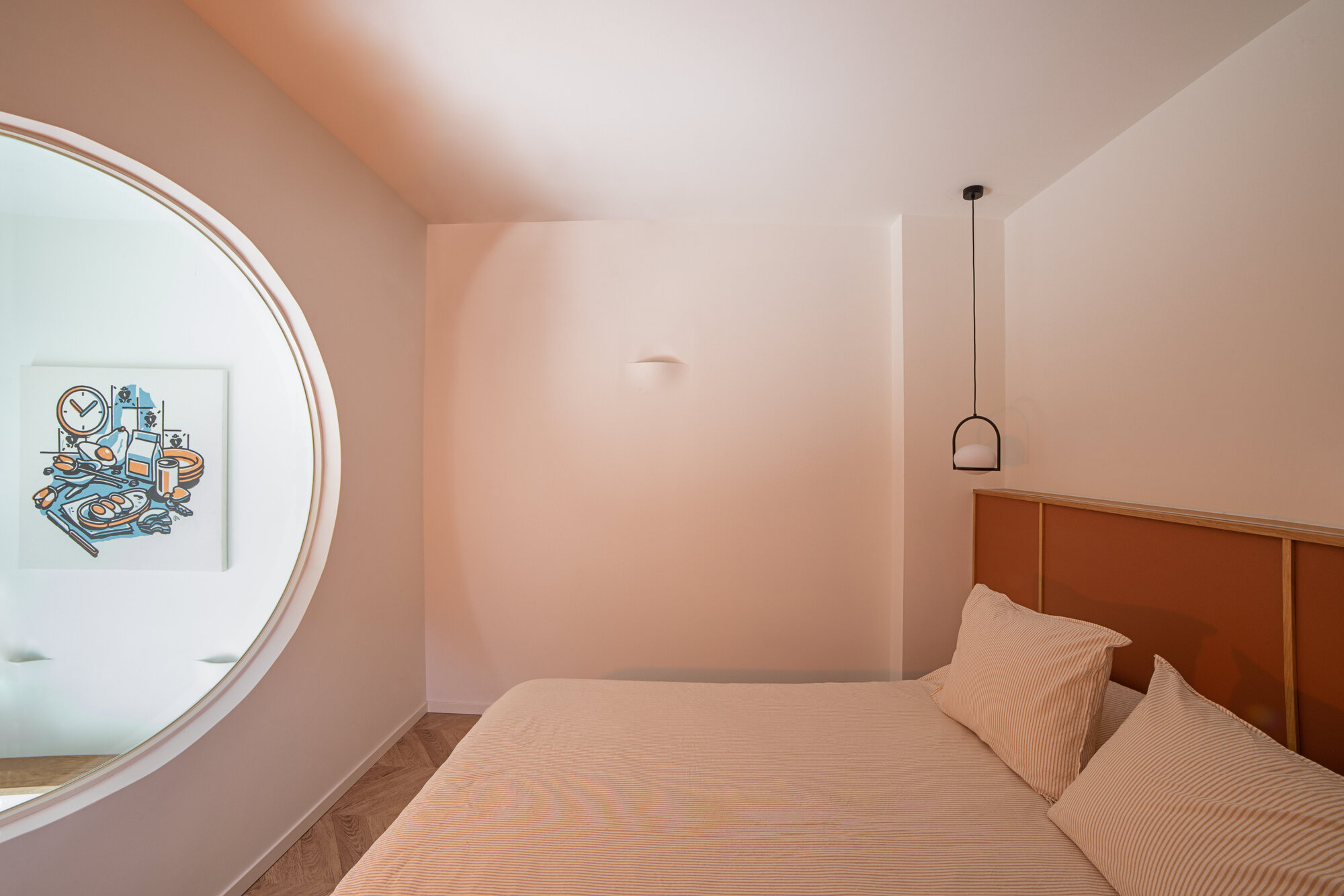 Bedroom seen past a round window opening onto an artwork, with a terracotta headboard, striped pillows and a black pendant lamp in warm light