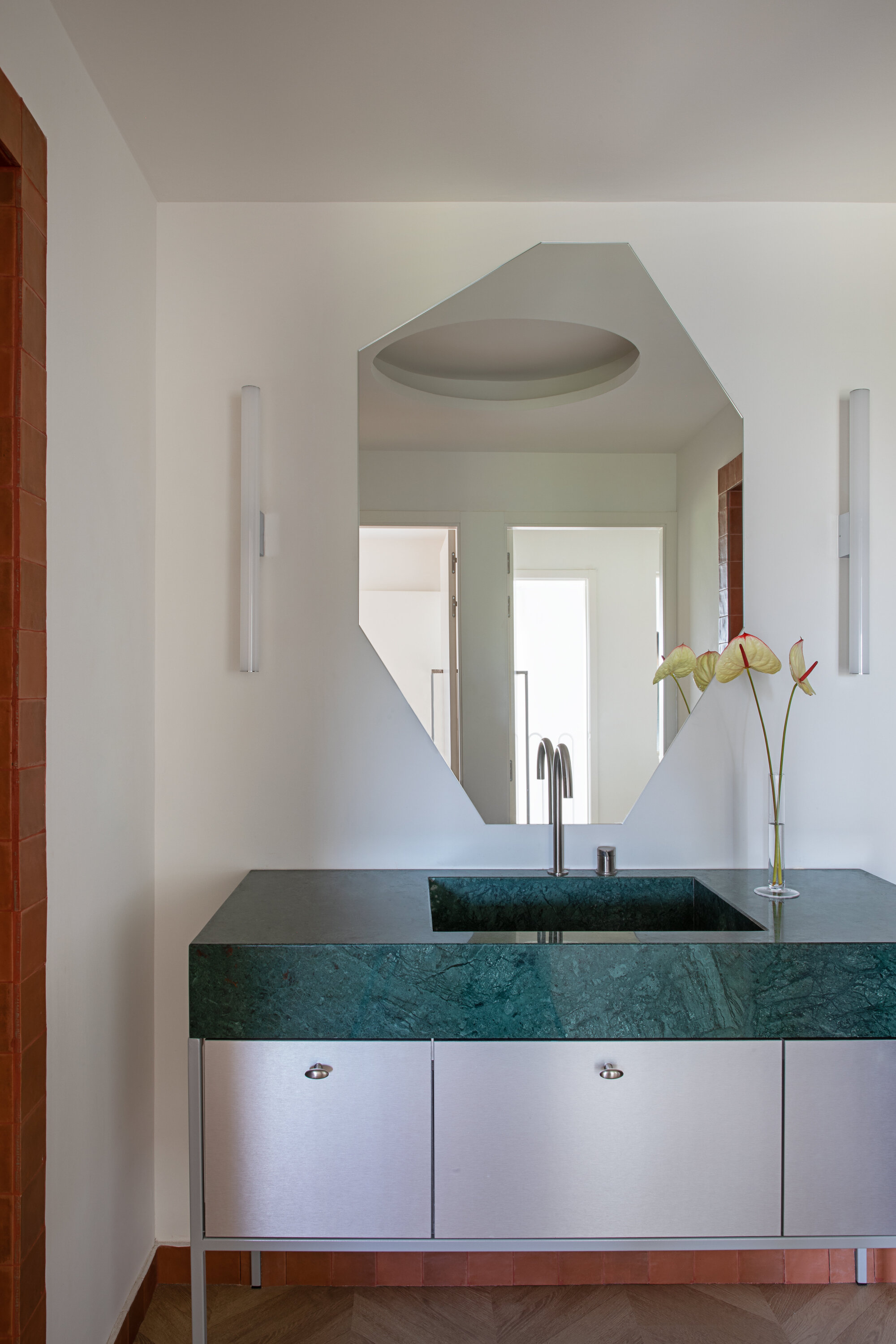 Green marble vanity with brushed steel cabinet doors beneath an angular mirror, flanked by tube wall lights and a vase of anthuriums