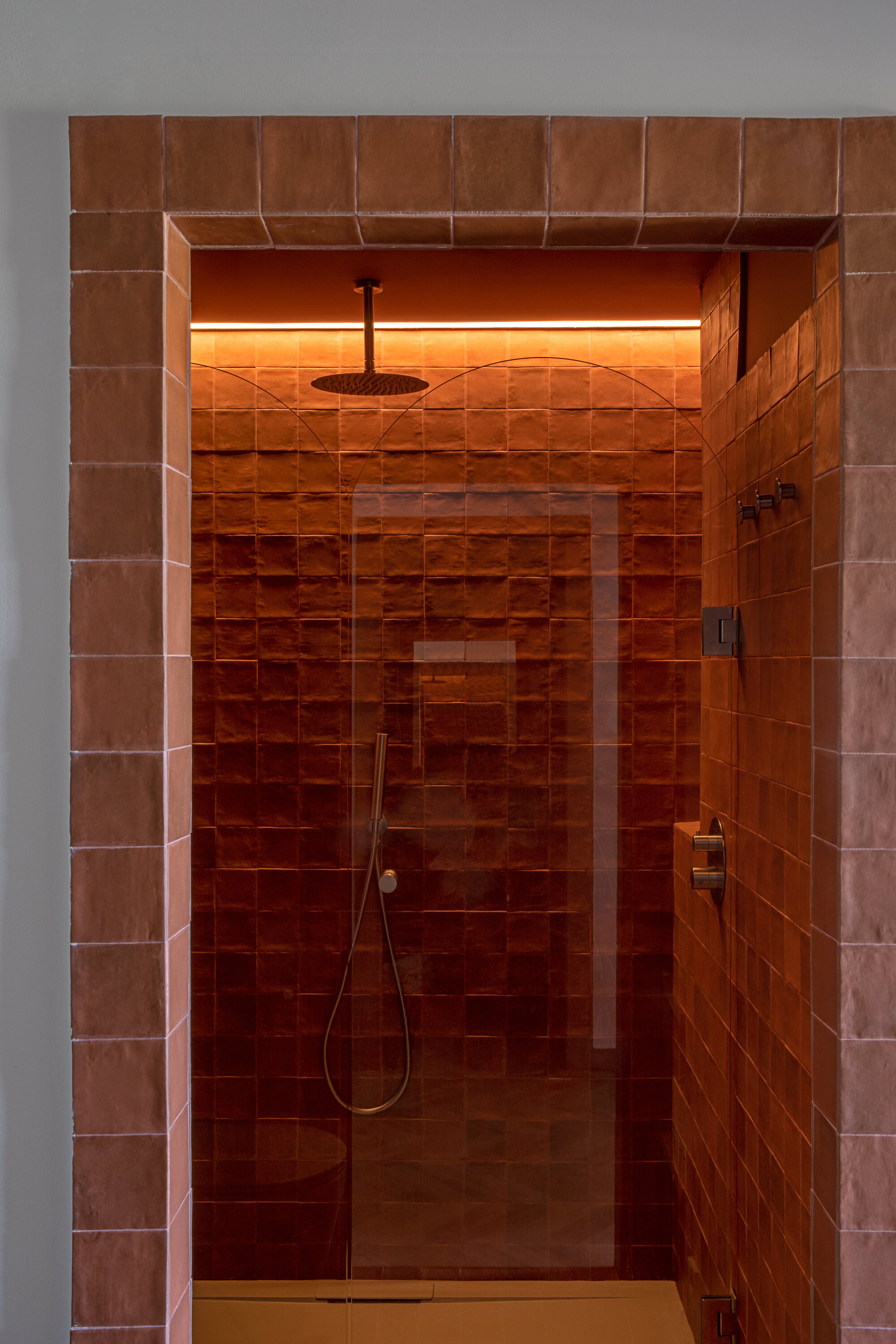 Shower alcove lined in terracotta glazed tiles with a ceiling rain head, hand shower and a warm strip of light above the glass