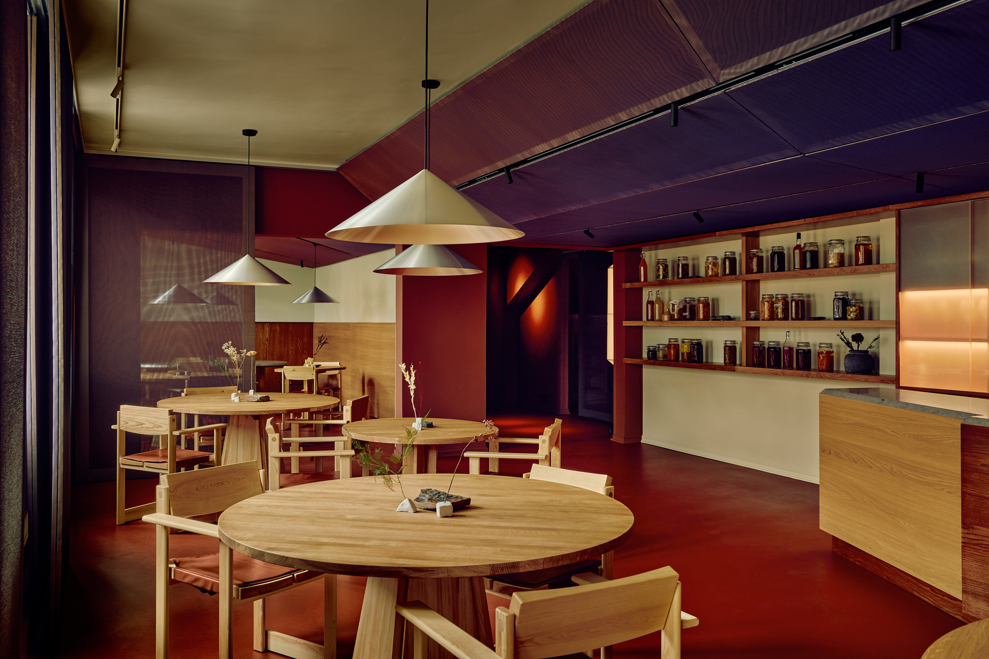 Dining room with round oak tables, conical pendant lamps and a shelf of preserve jars against red walls and floor