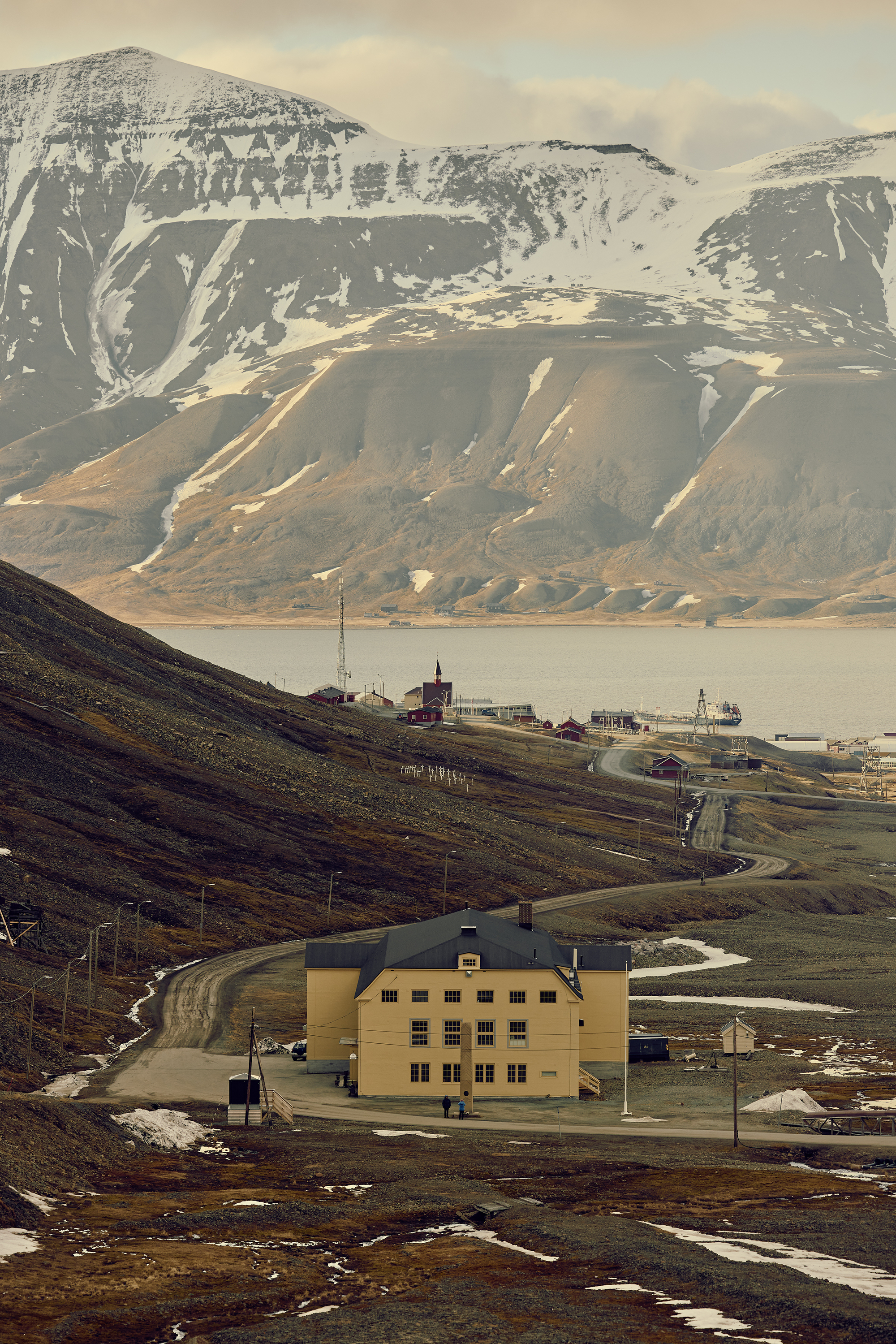 Yellow gabled building on a gravel road below snow-streaked mountains, with a fjord and small settlement behind in low light