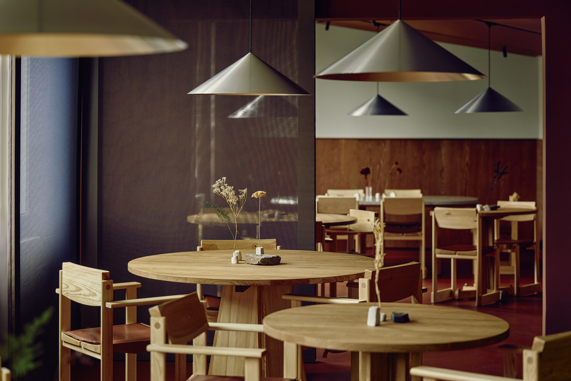 Round oak tables with dried flower stems under conical metal pendants, seen past a dark mesh curtain in dim light