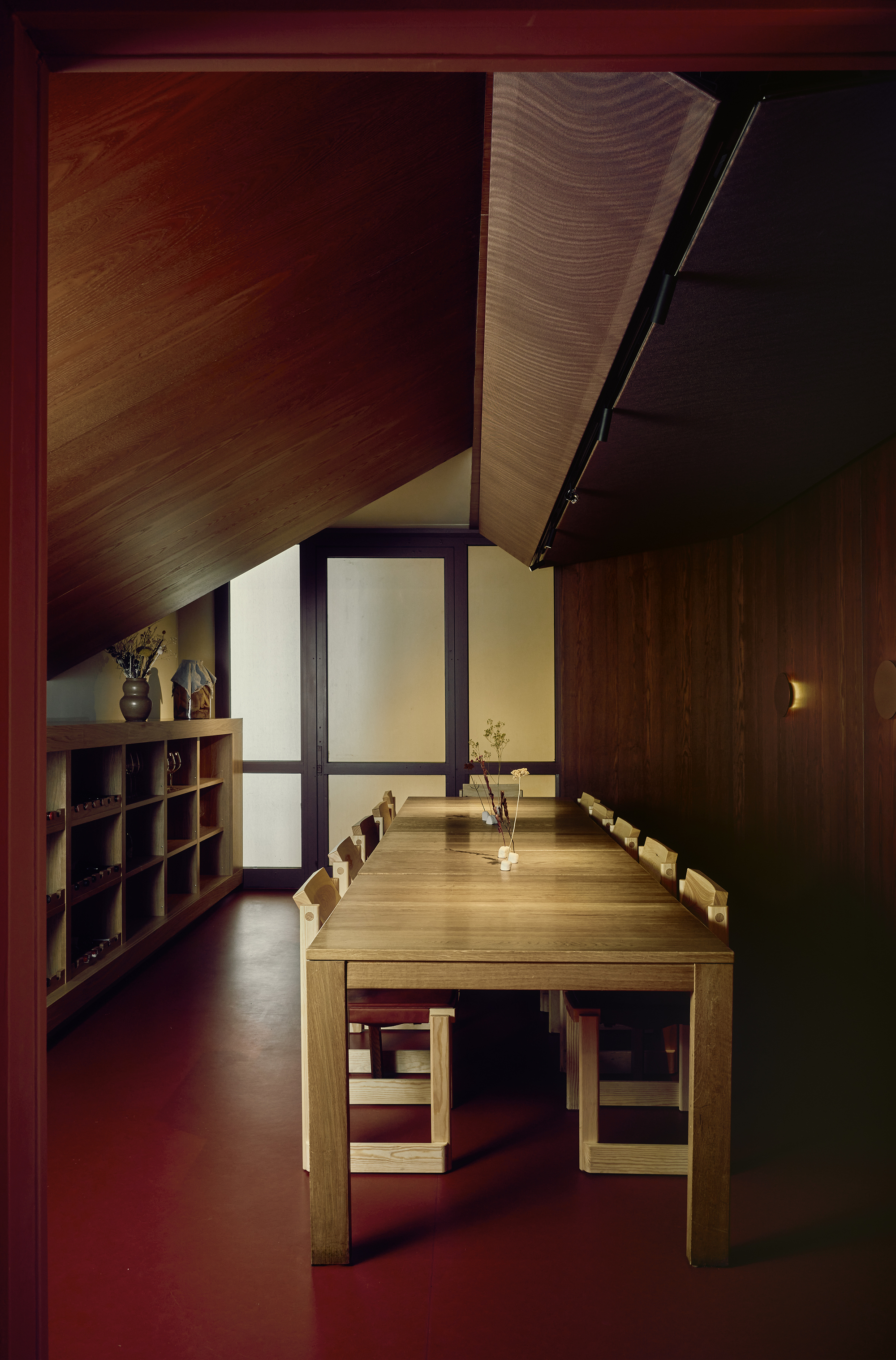 Long oak table and chairs beneath a sloped timber ceiling, with open shelving and frosted glass doors at the far end