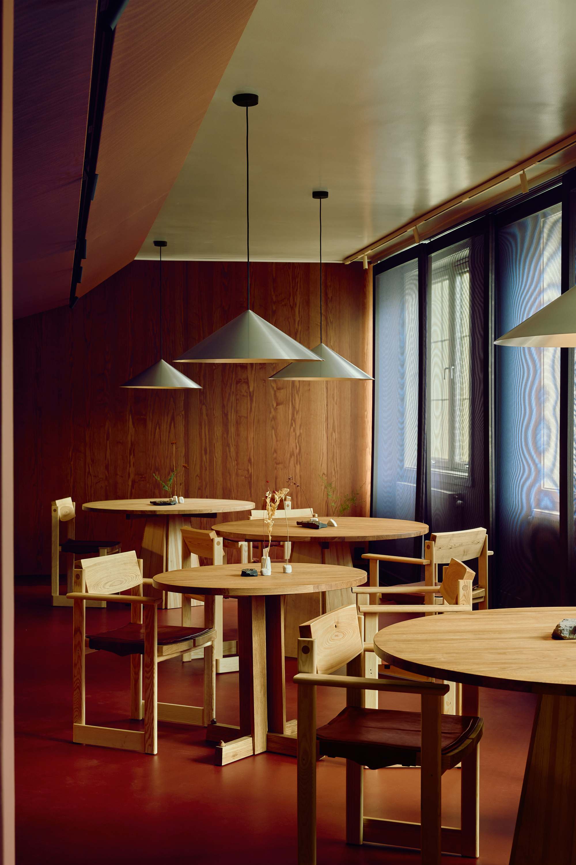 Oak tables and armchairs on a red floor beneath cone pendants, with sheer dark curtains over the tall windows