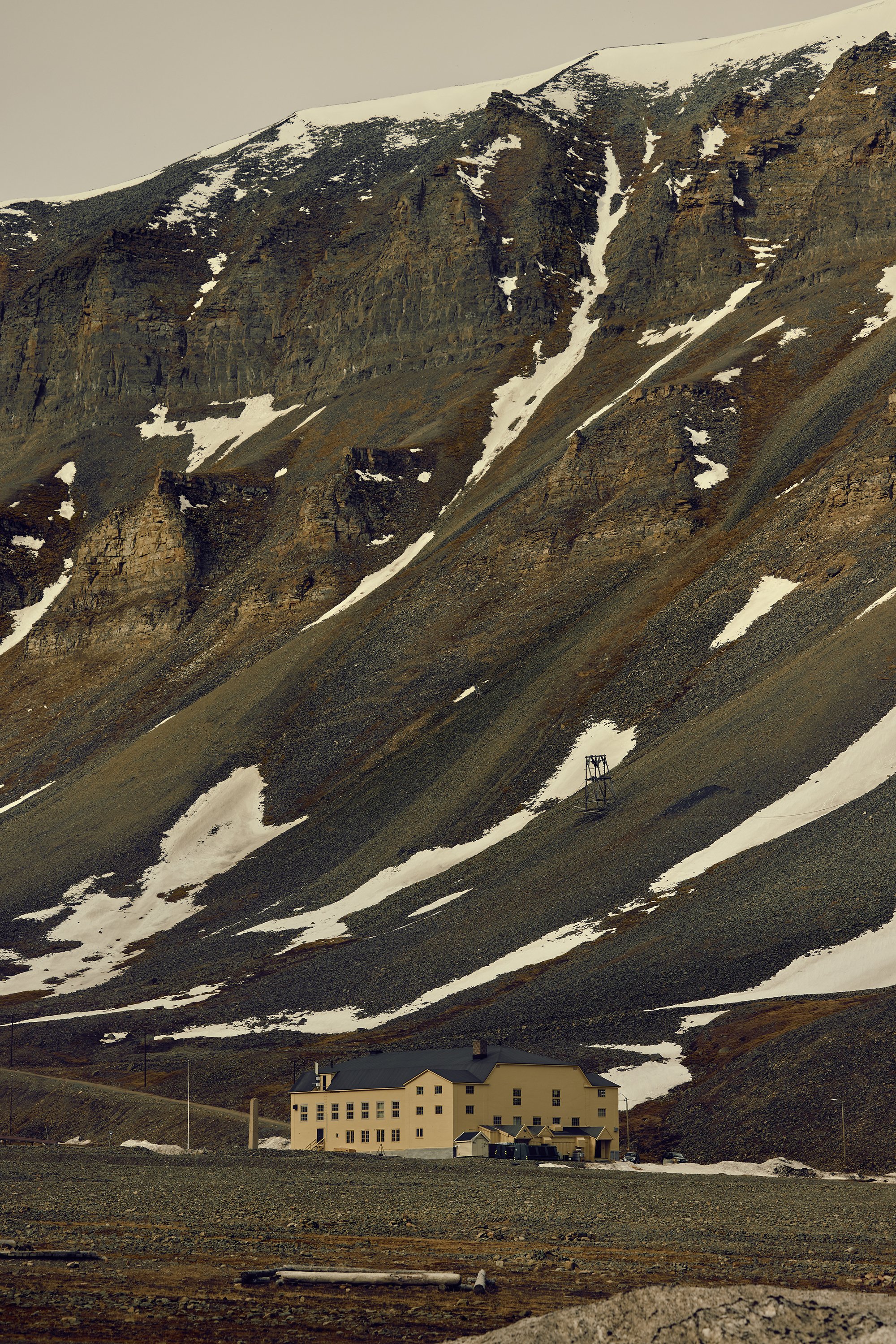 Yellow multi-storey building with a dark roof at the foot of a steep scree slope streaked with patchy snow