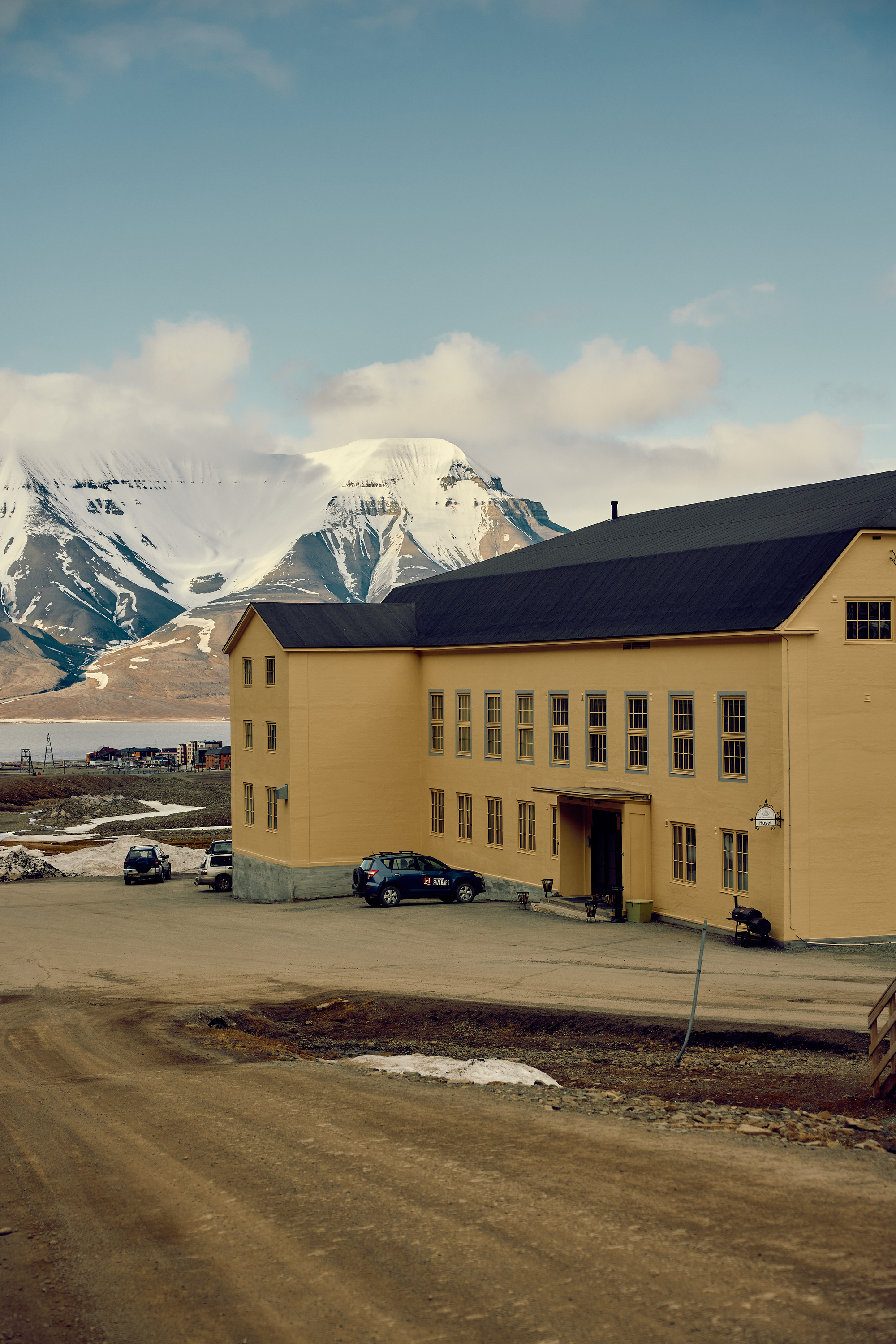 Yellow timber-clad building with dark pitched roof and entrance canopy, snowy peak and cloud banks behind it