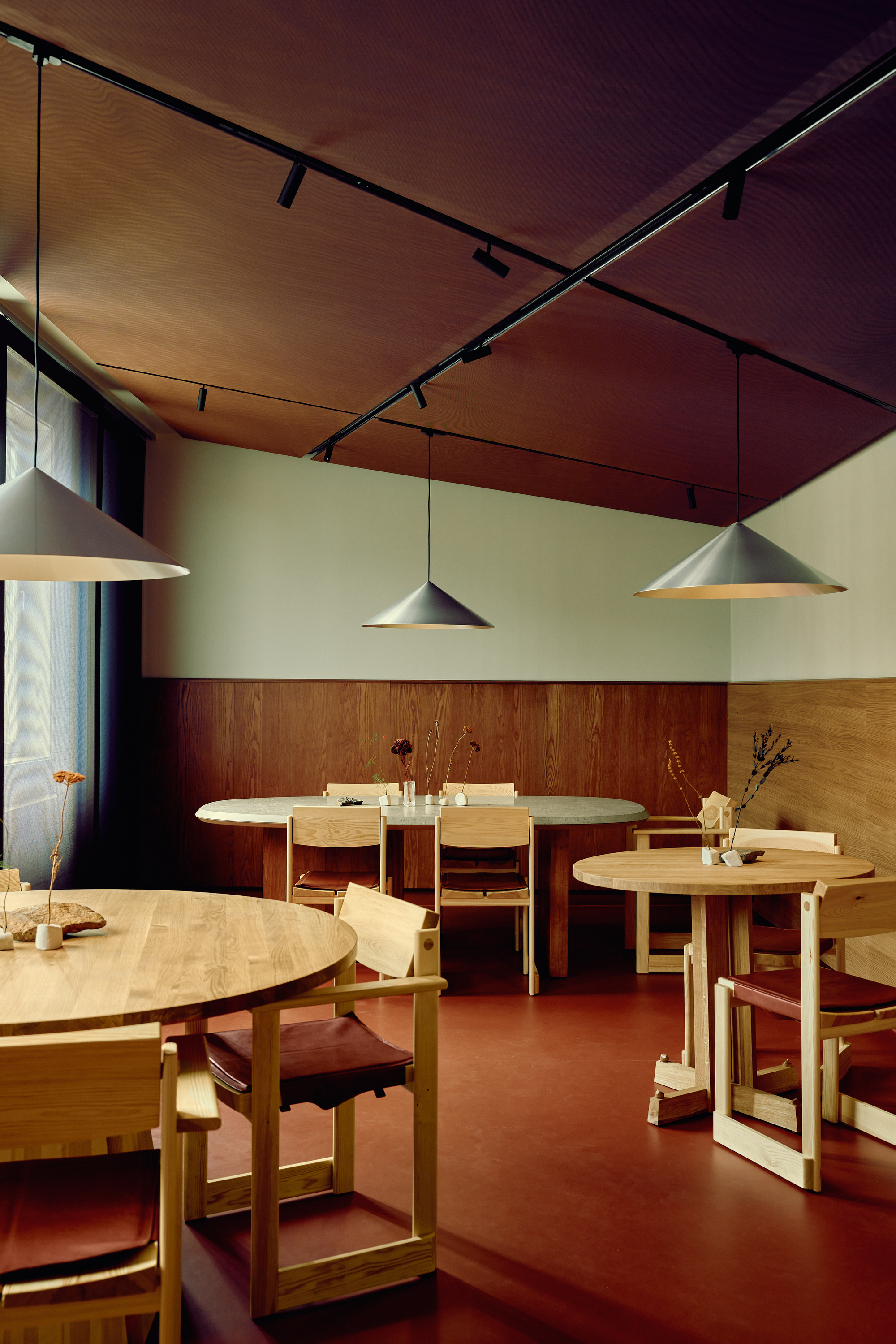 Dining room with round oak tables, wooden armchairs and conical pendant lamps under a dark track-lit ceiling