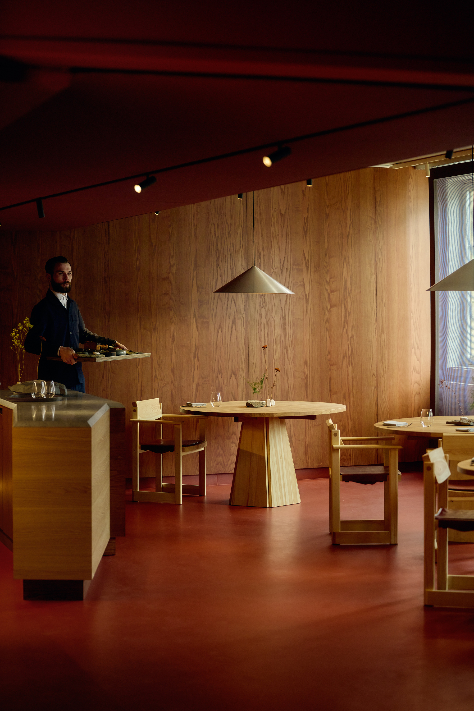 Server carrying a tray past a stone-topped oak counter, with round tables on a rust-red floor in low light