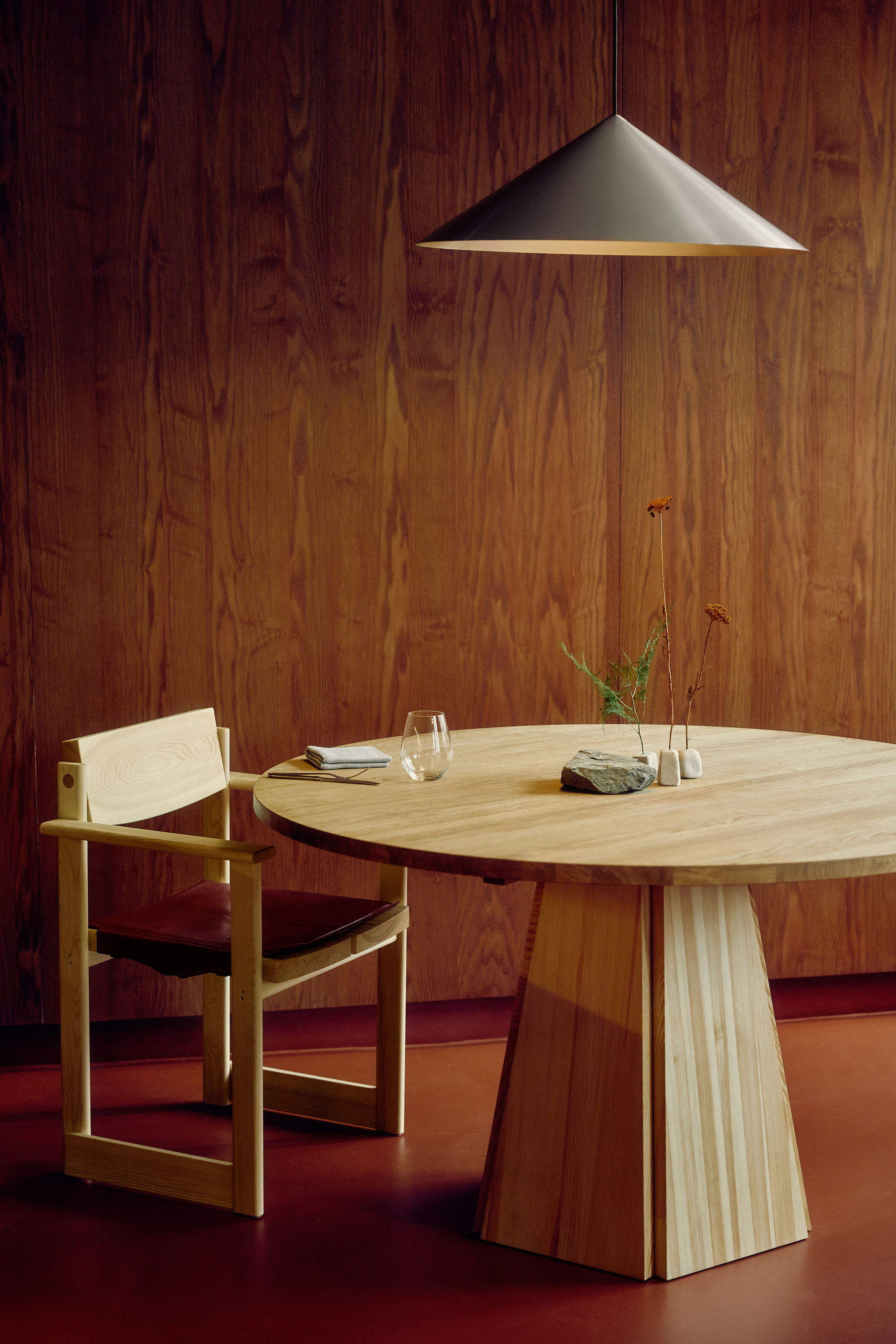 Round oak table with tapered pedestal base and leather-seated armchair beneath a conical pendant, set against grained wood paneling