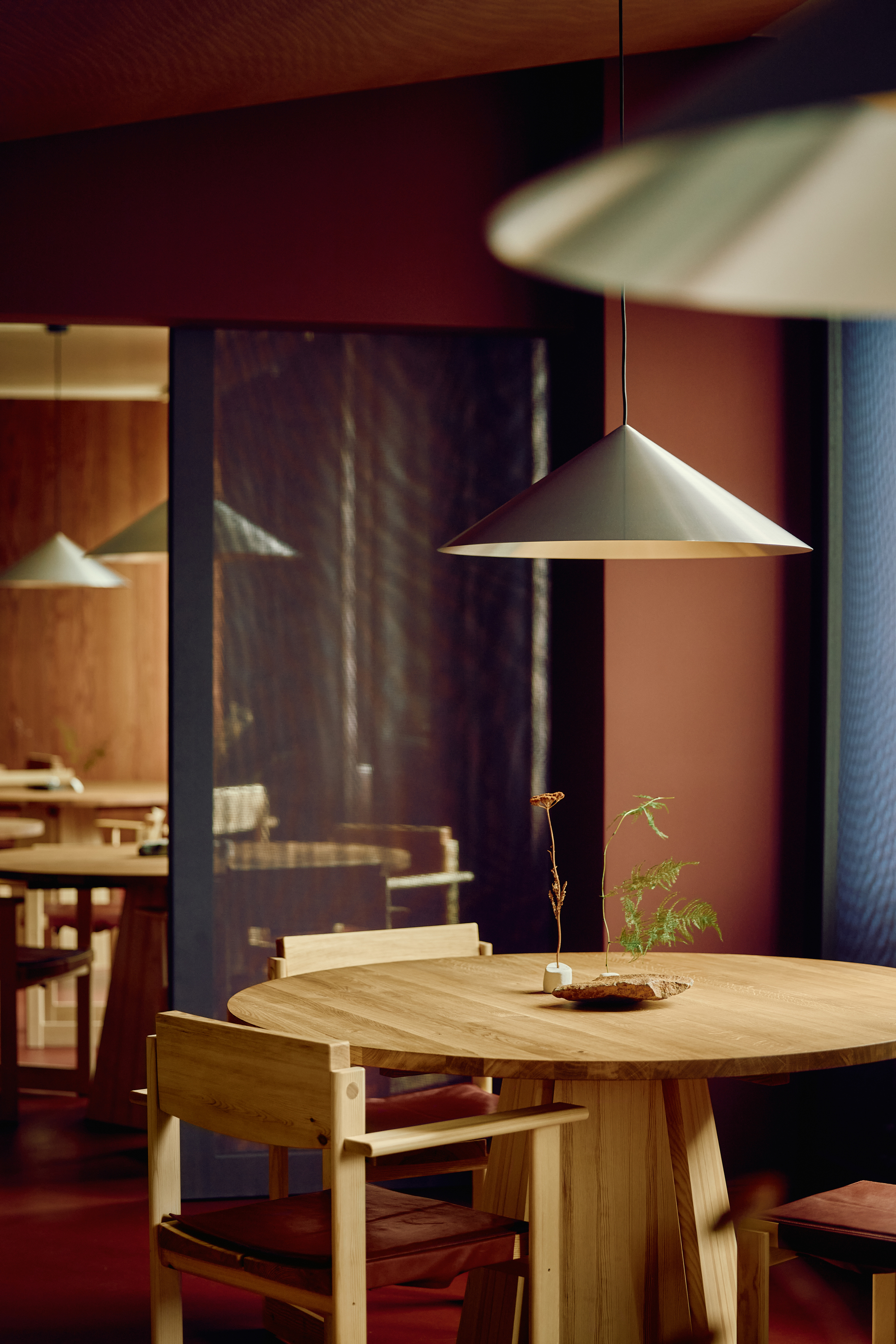 Round oak dining table with a dried-stem arrangement beneath a conical pendant lamp, set against terracotta walls in low warm light