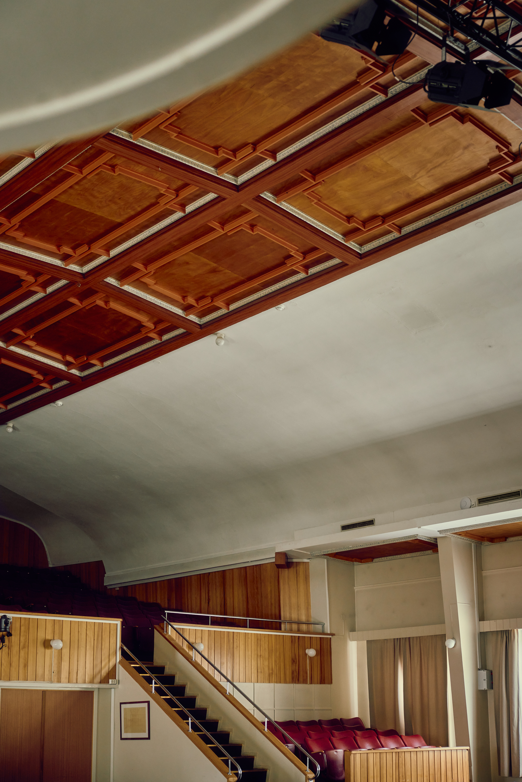 Auditorium with a coffered timber ceiling, red seating and a side staircase, lit by soft daylight from curtained windows