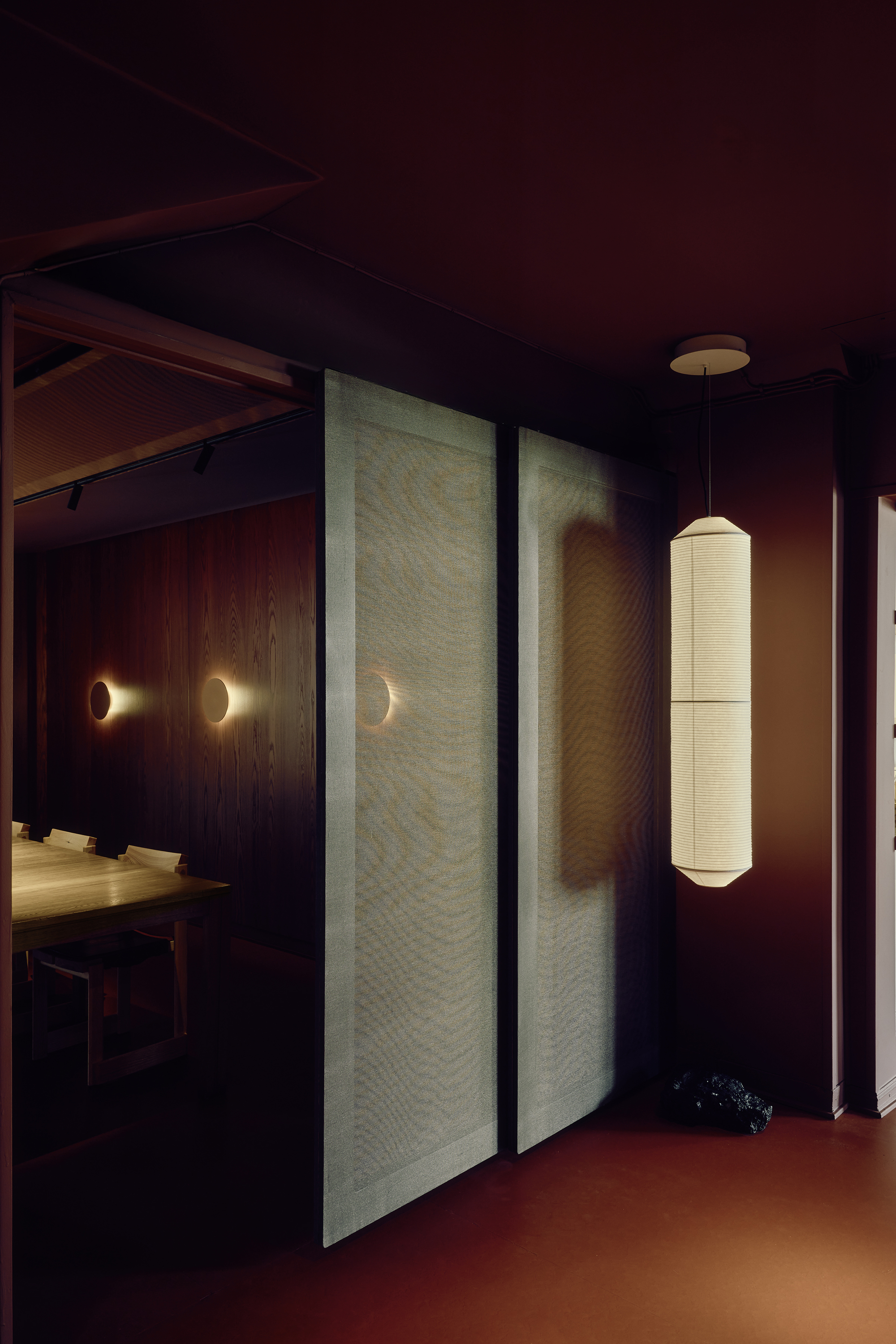 Sliding fabric-panel doors beside a tall paper pendant lamp, with wall sconces glowing in a dark red-brown room