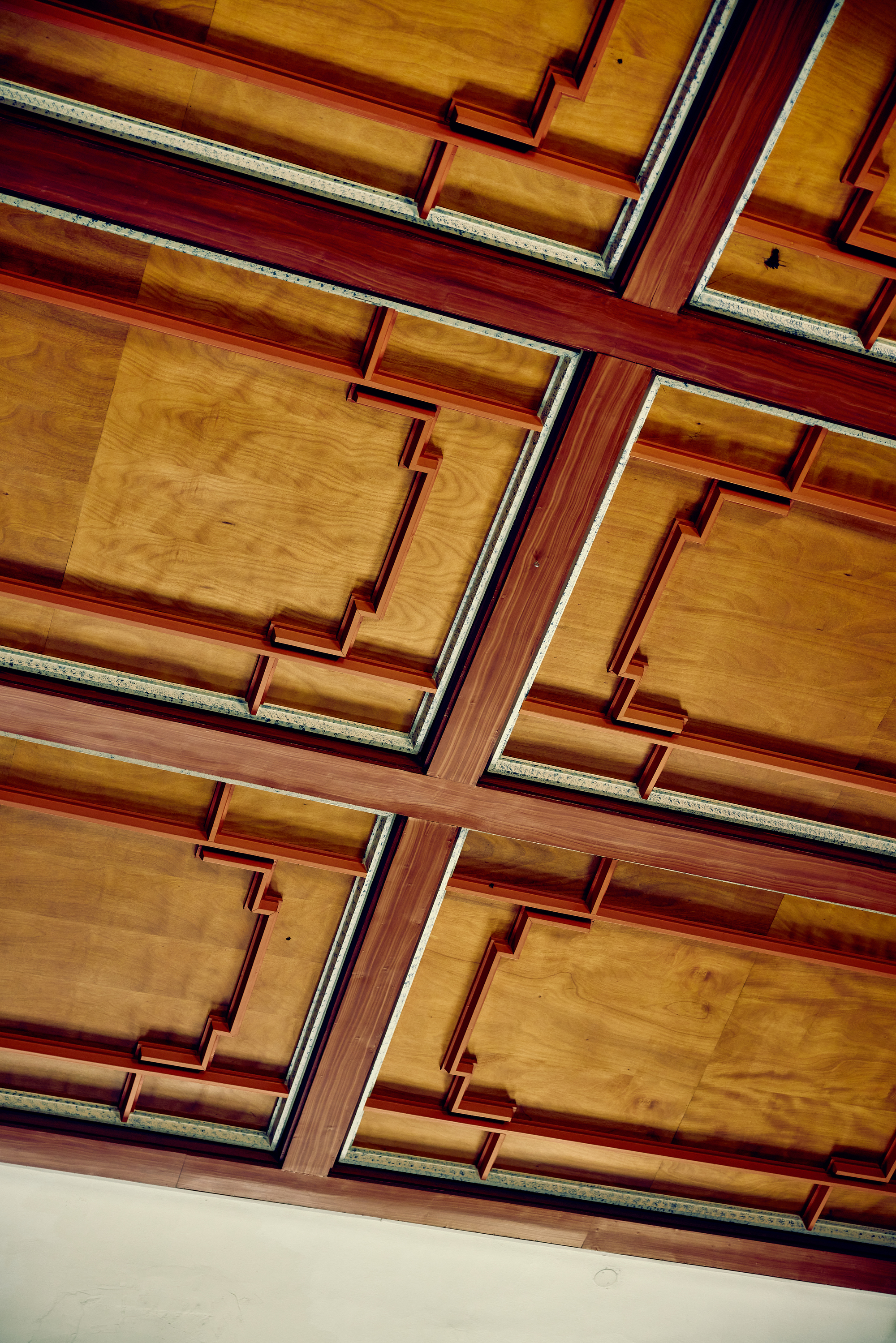 Looking up at a coffered plywood ceiling with red-stained timber beams and stepped trim in each panel, under even daylight