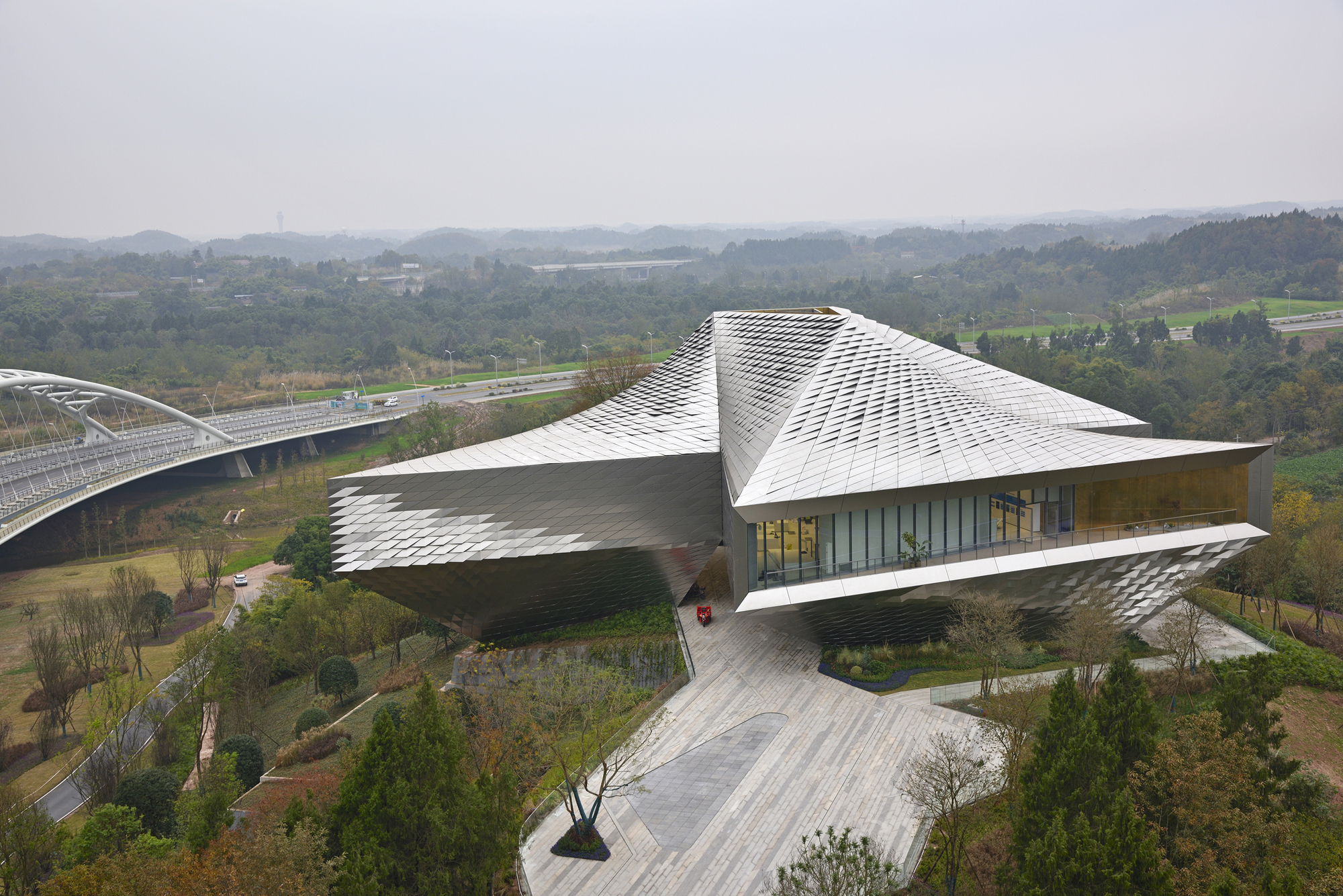 Aerial view of the faceted metal-clad roof and glazed upper floor beside a highway bridge, under hazy grey skies