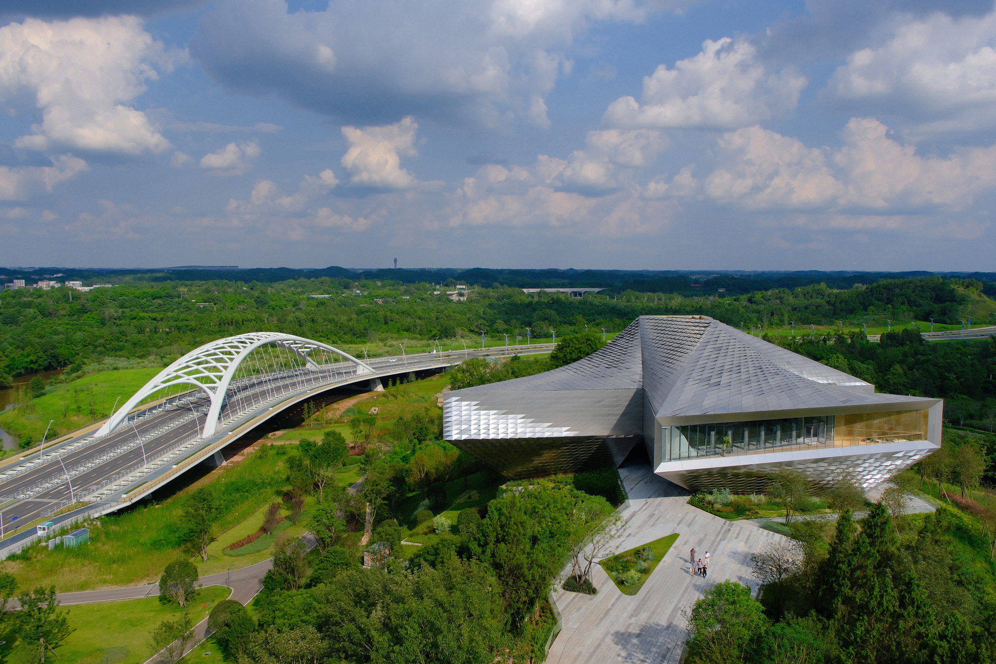 Drone view of the angular scaled roof and paved forecourt next to a white arched bridge under cumulus clouds
