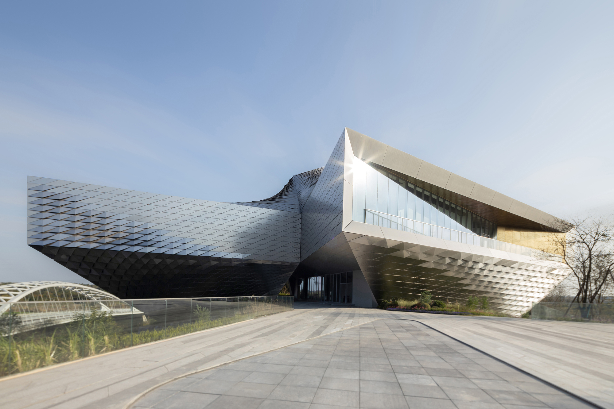 Low view of the cantilevered faceted facade and glazed corner over a paved plaza in soft late light