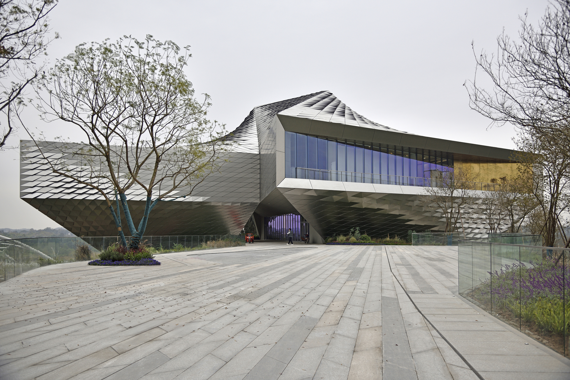 Entrance front with blue glazing and overhanging scaled metal volumes behind a plaza of long stone pavers and a bare tree