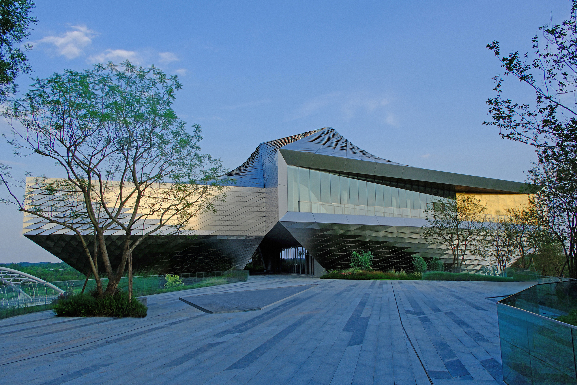 Entrance plaza of striped stone paving leading to the angular metal-clad volume at dusk, with a slim tree in the foreground