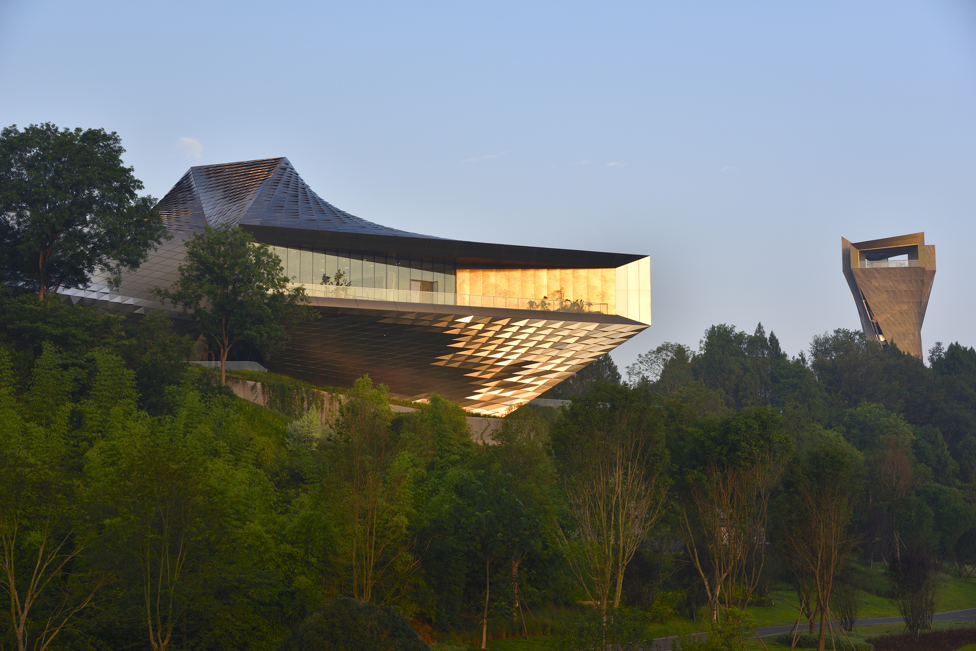 Cantilevered box with a gold-lit end wall and diamond-patterned underside, with a tower rising beyond the trees in low evening sun