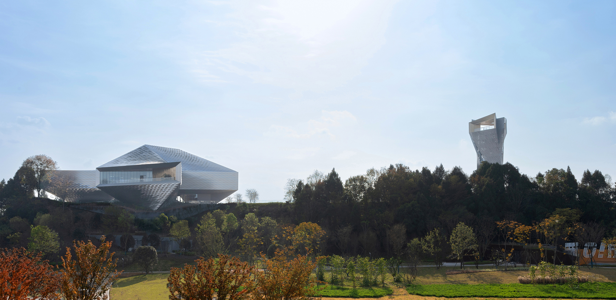 Wide view of the faceted metal building and a distant lookout tower above a tree line in hazy autumn light