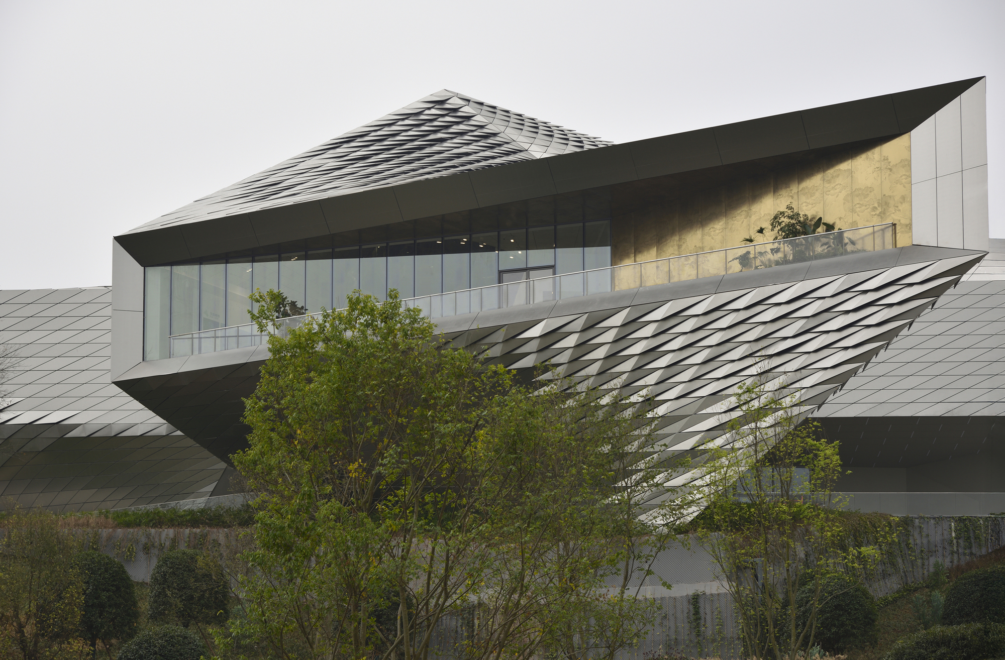 Cantilevered upper volume clad in faceted metal scales, with a glazed band and terrace rail behind leafy trees under an overcast sky