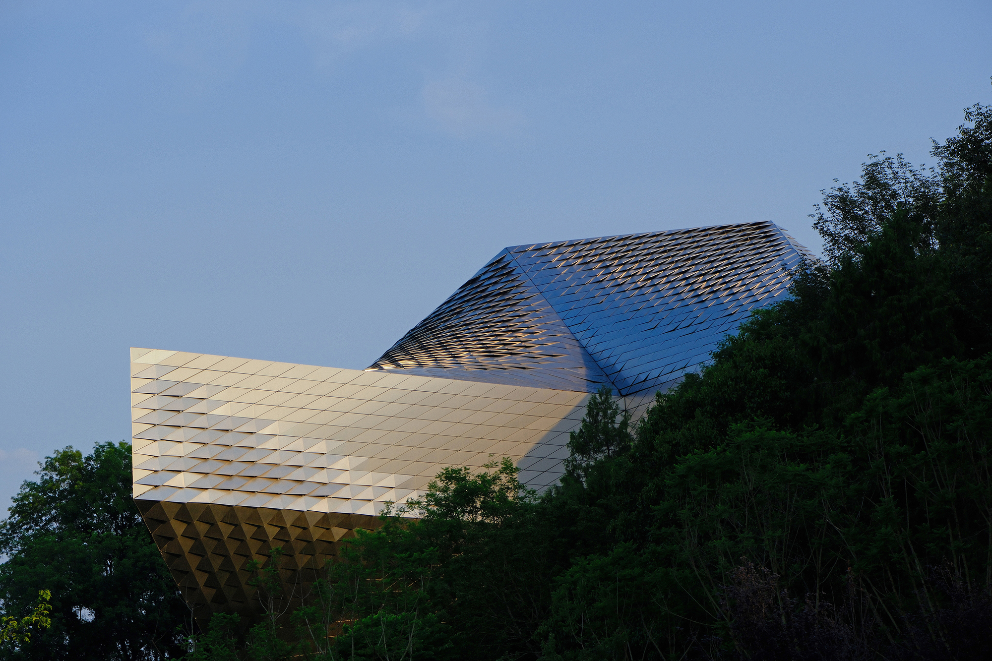 Overhanging faceted metal volume with a gold-toned underside and pyramidal roof rising above dark treetops at dusk
