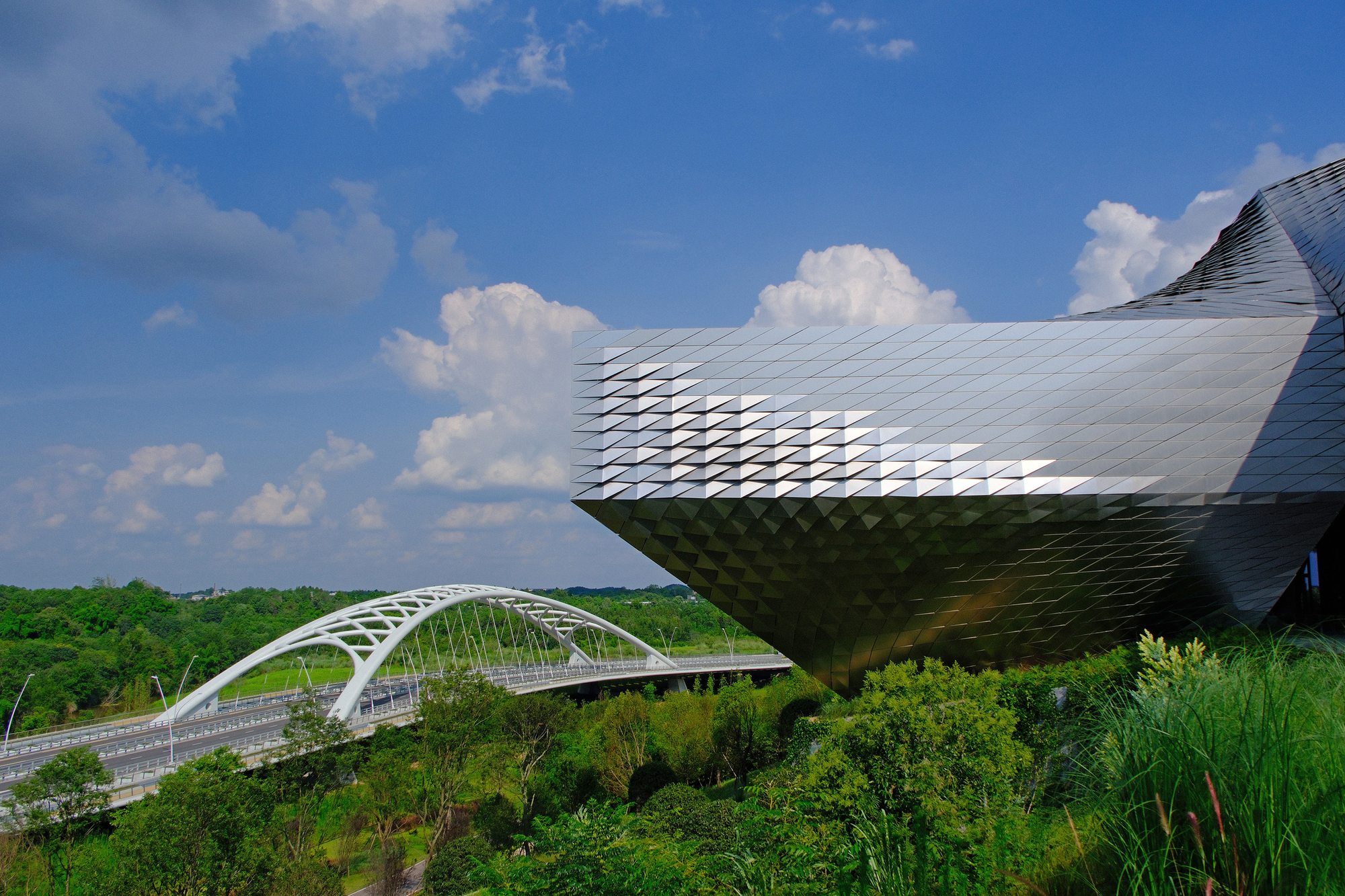 Cantilevered metal-tiled mass with a faceted underside beside a white arched bridge, under towering cumulus clouds