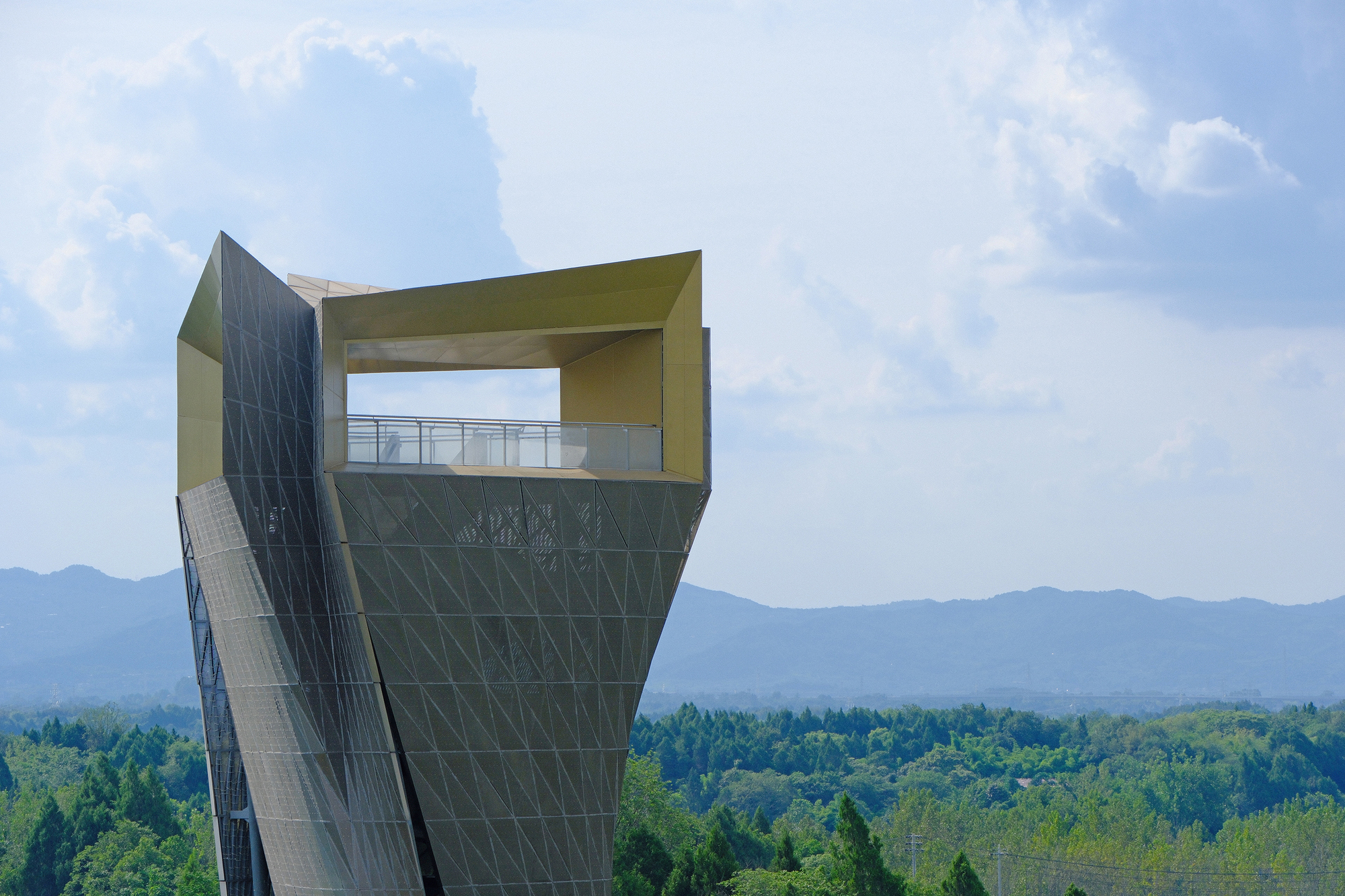 Twisting tower with a diagonal lattice skin and a gold-lined open viewing deck, set against distant hazy mountains