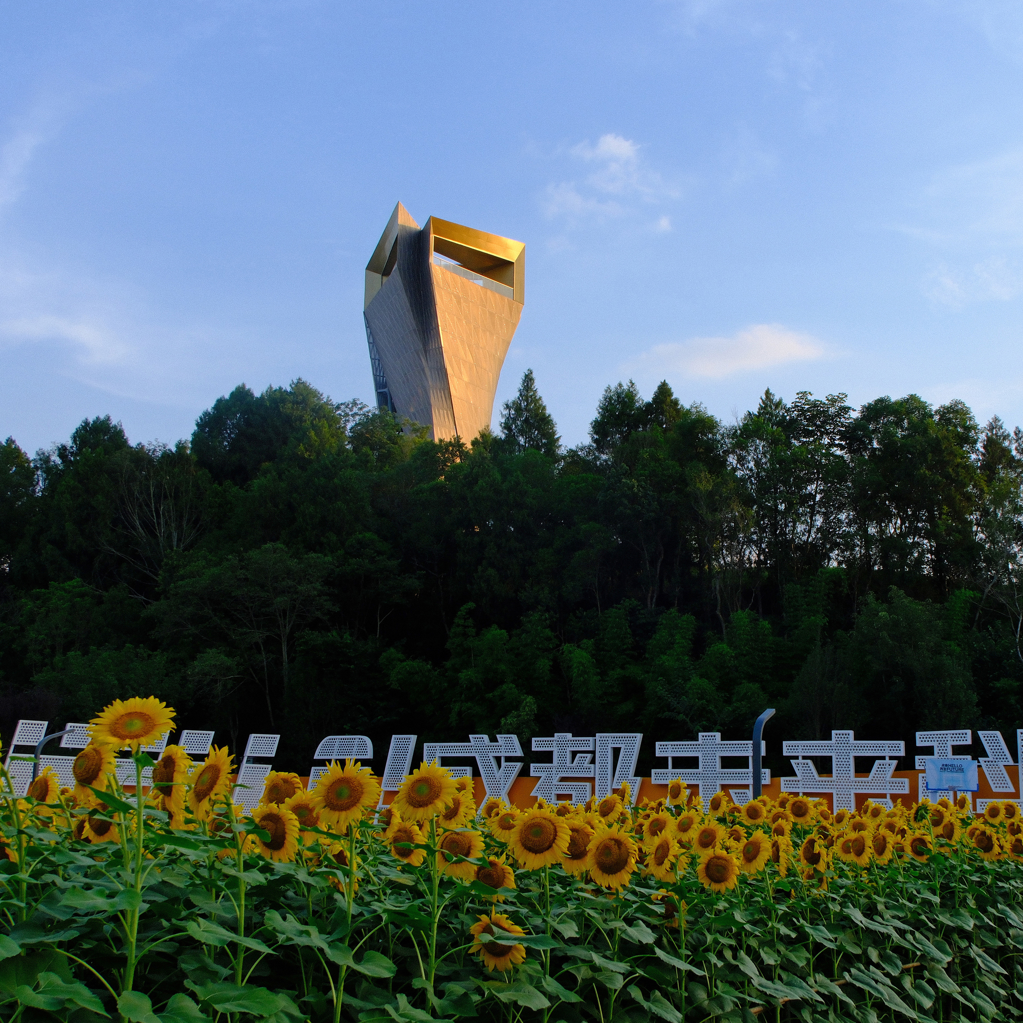 Gold-clad tower rising above a forested ridge, with a field of sunflowers and white lettering in the foreground