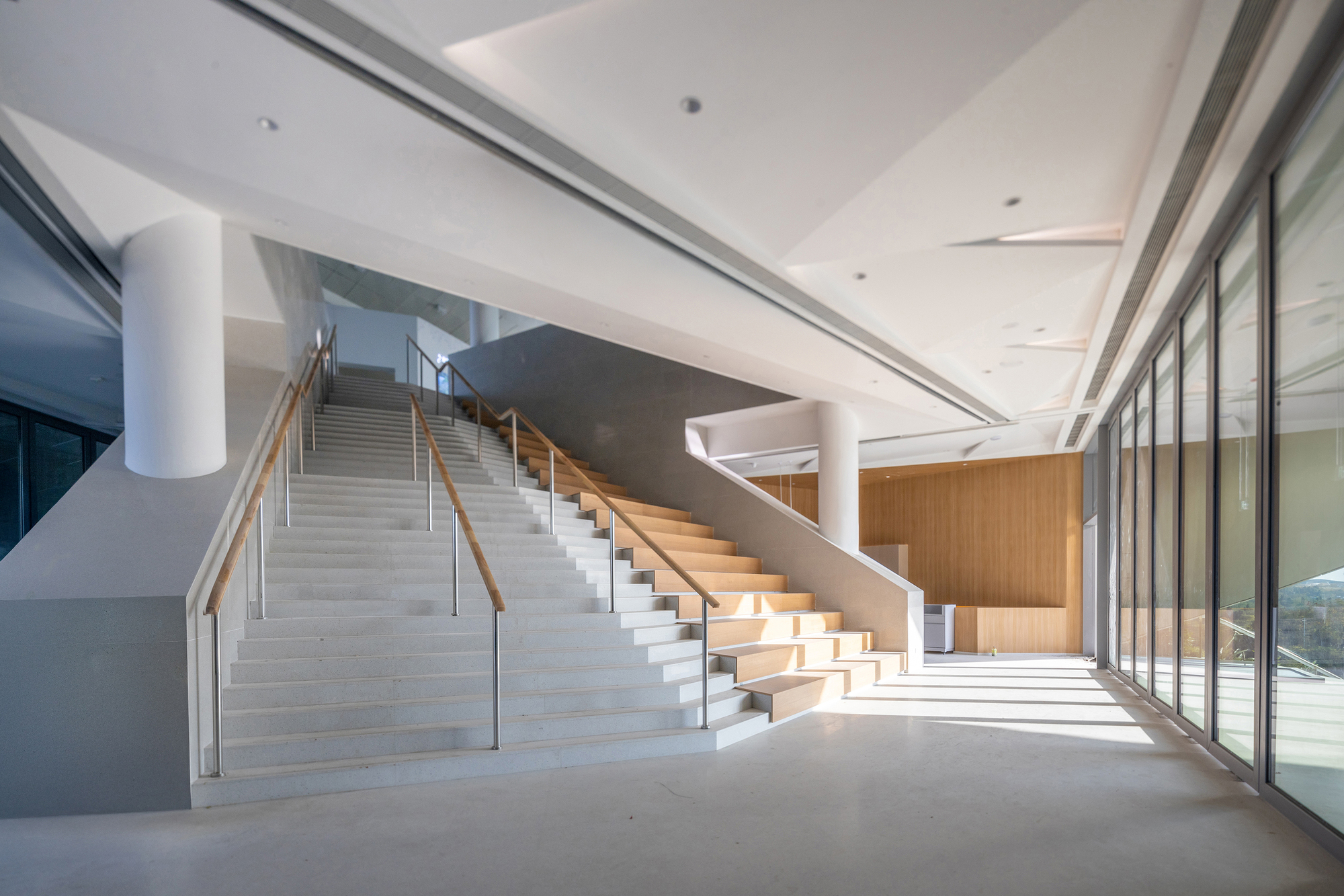 Broad white stair with timber-capped handrails and round columns in a hall where sunlight streams through glazing onto the floor