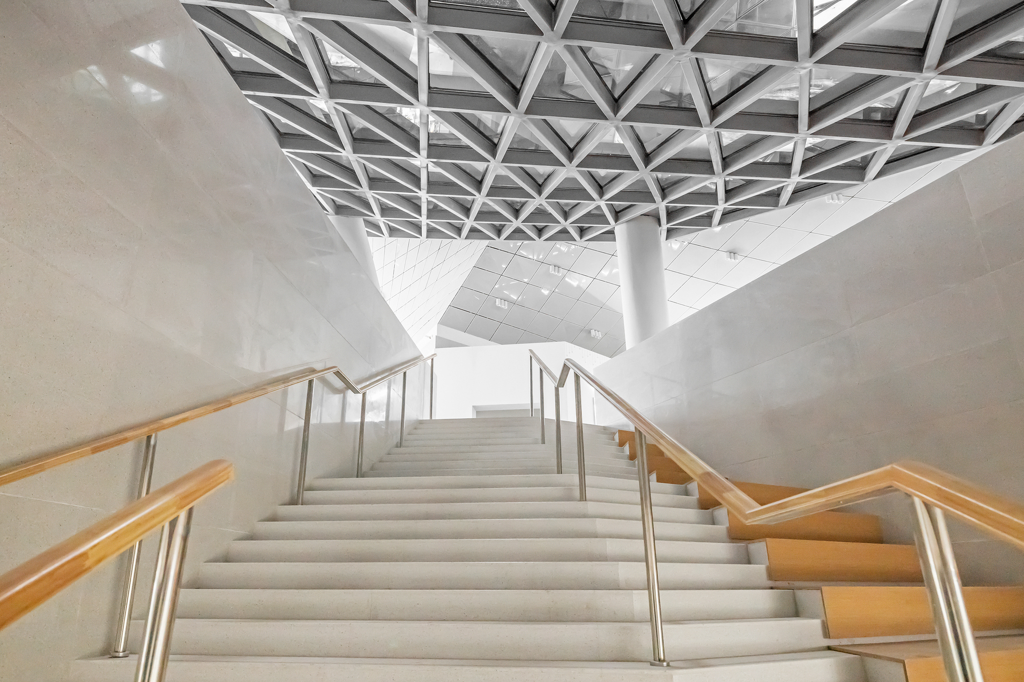 Looking up a white stair with timber treads toward a triangulated steel lattice ceiling and a round column