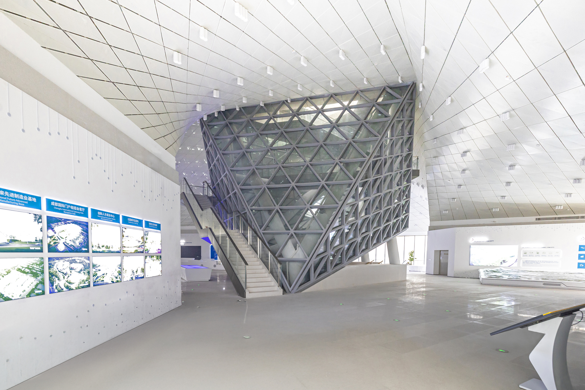 Faceted steel-and-glass volume beside a stair in an exhibition hall, with wall-mounted display panels under a folded white ceiling