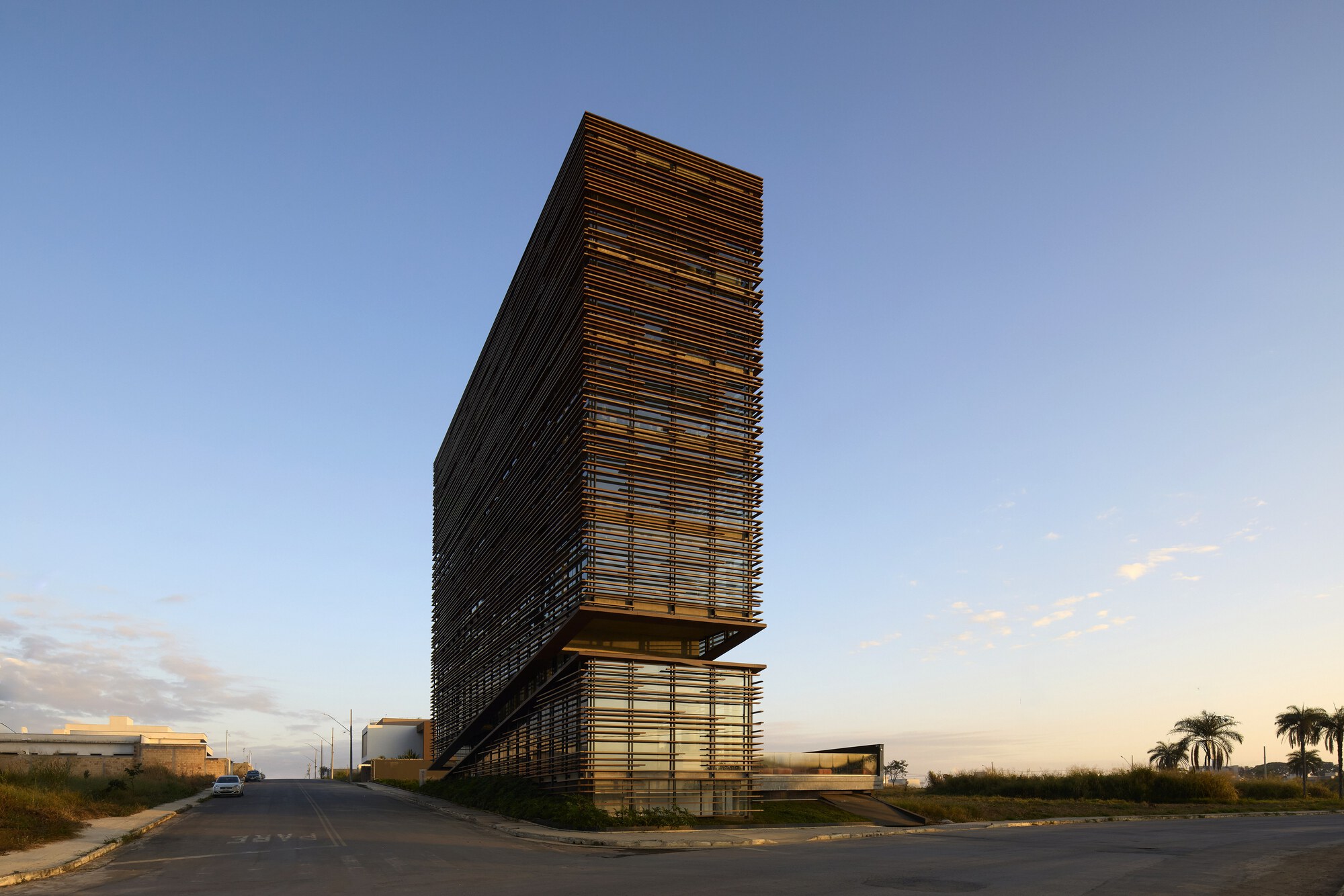 Corner view of the tilted timber-louvered tower, its upper block cantilevering over a glazed base against a clear dusk sky