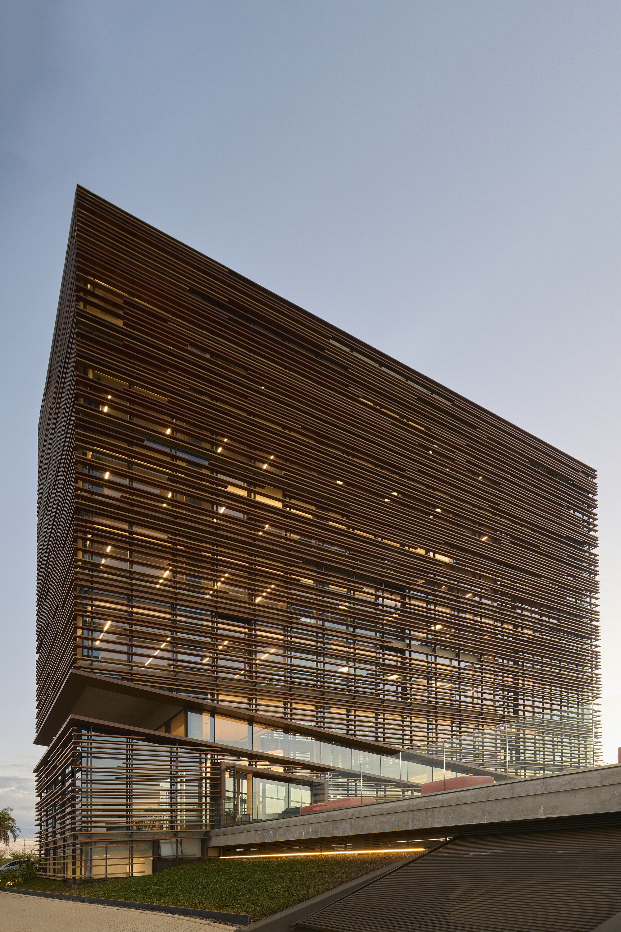 Low angle of the wooden louvered volume with small ceiling lights glowing behind the slats above a grassy slope and concrete wall