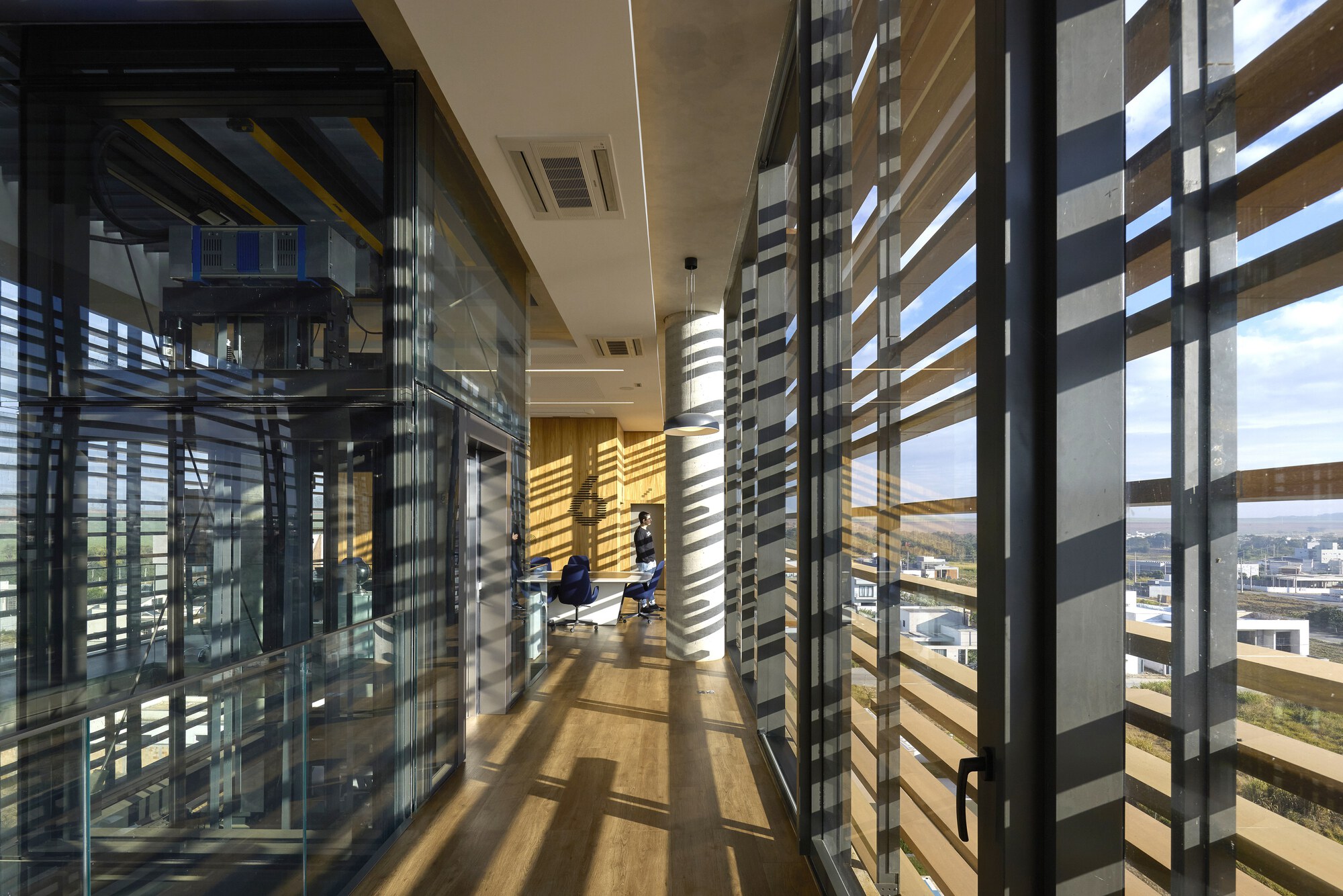 Interior corridor along the louvered glass wall, with striped sunlight across the timber floor and a glass lift shaft on the left